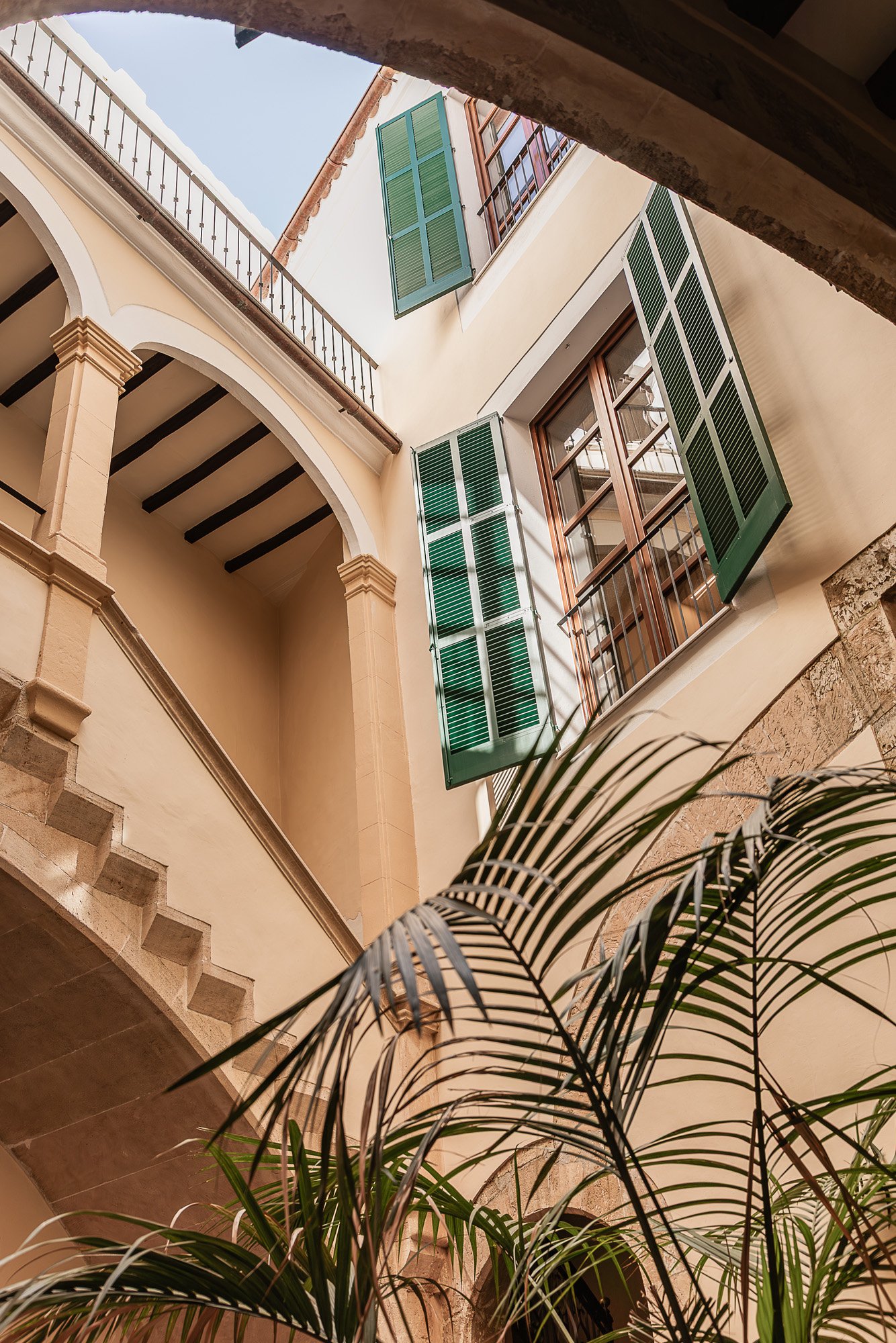 View of an interior courtyard with open green window shutters, an arched walkways, and a ceiling with exposed dark wooden beams.
