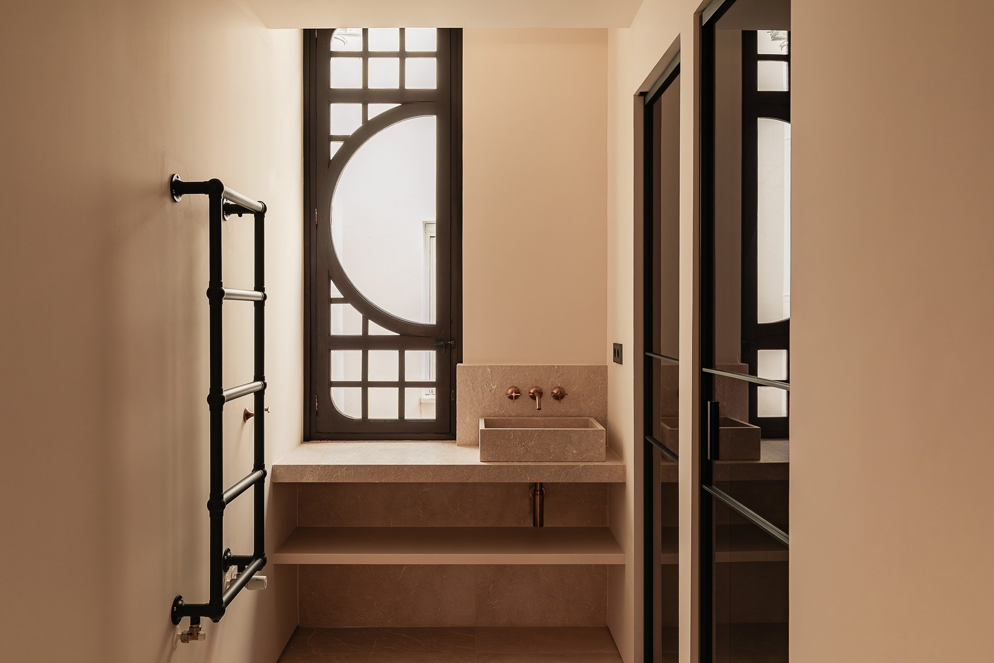Minimalist bathroom with a black window with geometric design, a small beige stone sink, a black towel rack, and a glass-fronted cabinet.