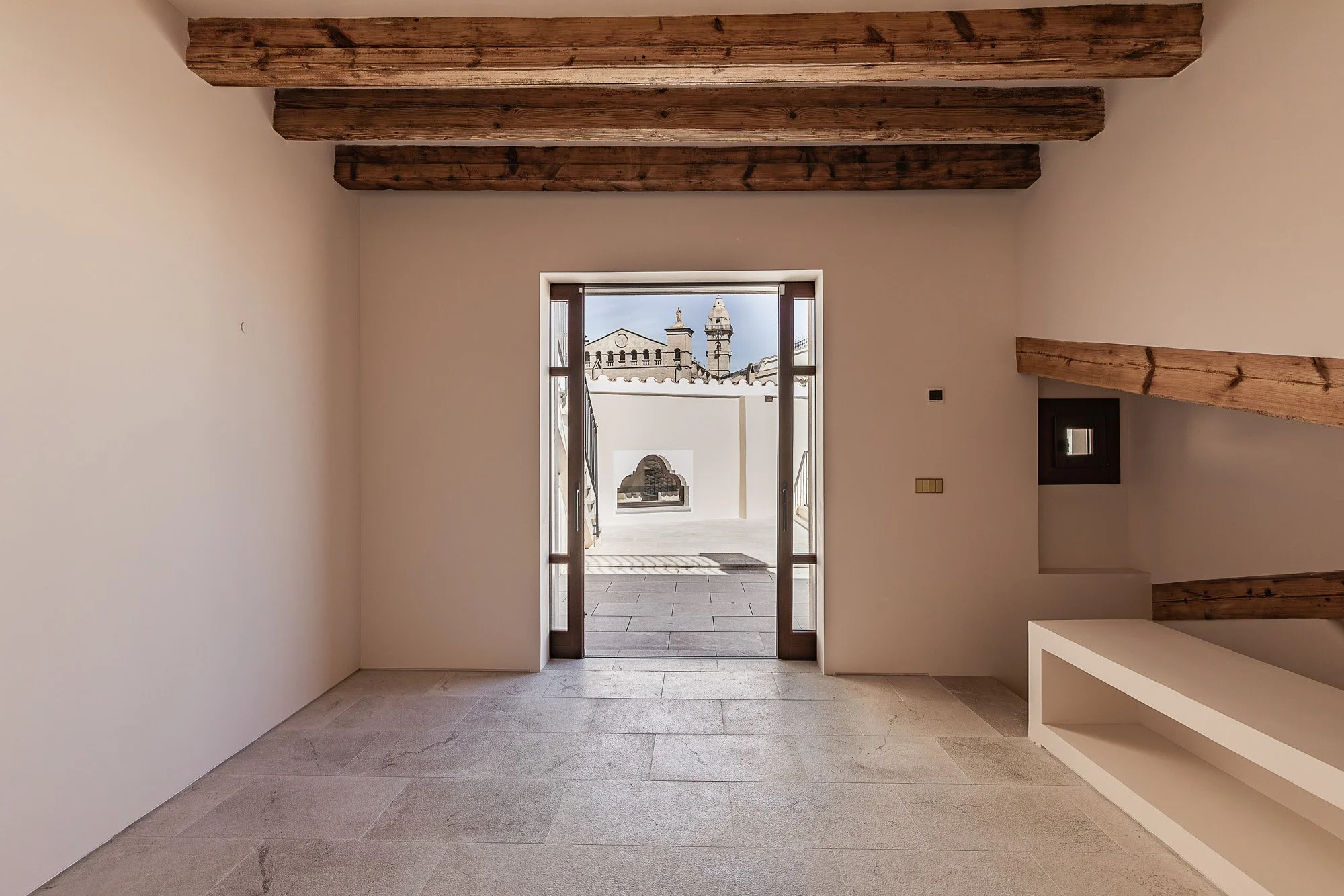 Empty room with beige walls, wooden ceiling beams, and a door opening to an outdoor terrace with a view of historic buildings and rooftops.