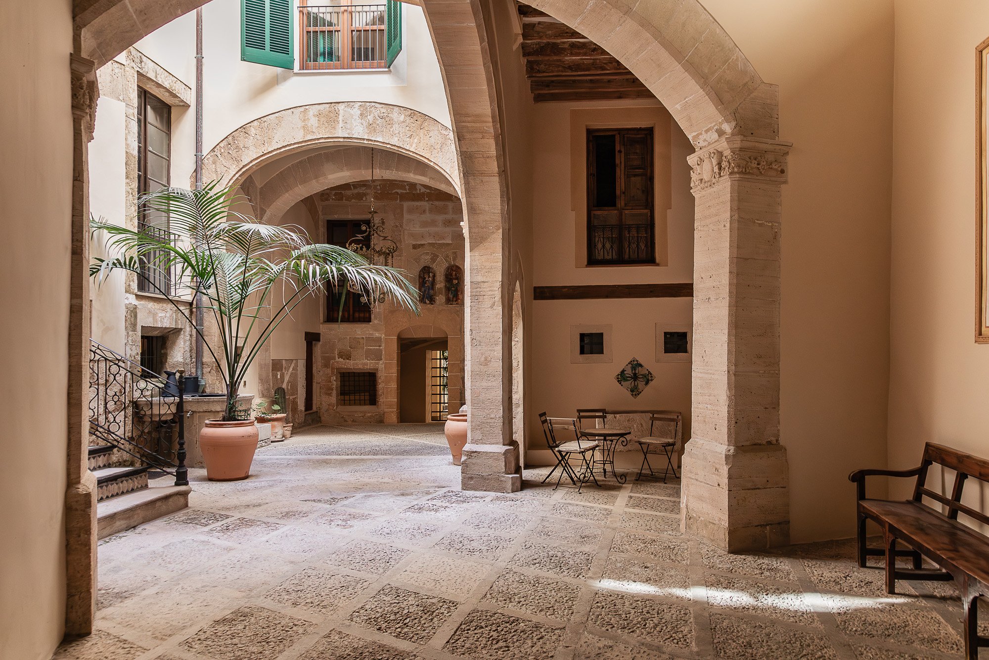 An interior courtyard of a historic building with stone arches, a potted plant, small tables and chairs, and a bench, with sunlight streaming in.