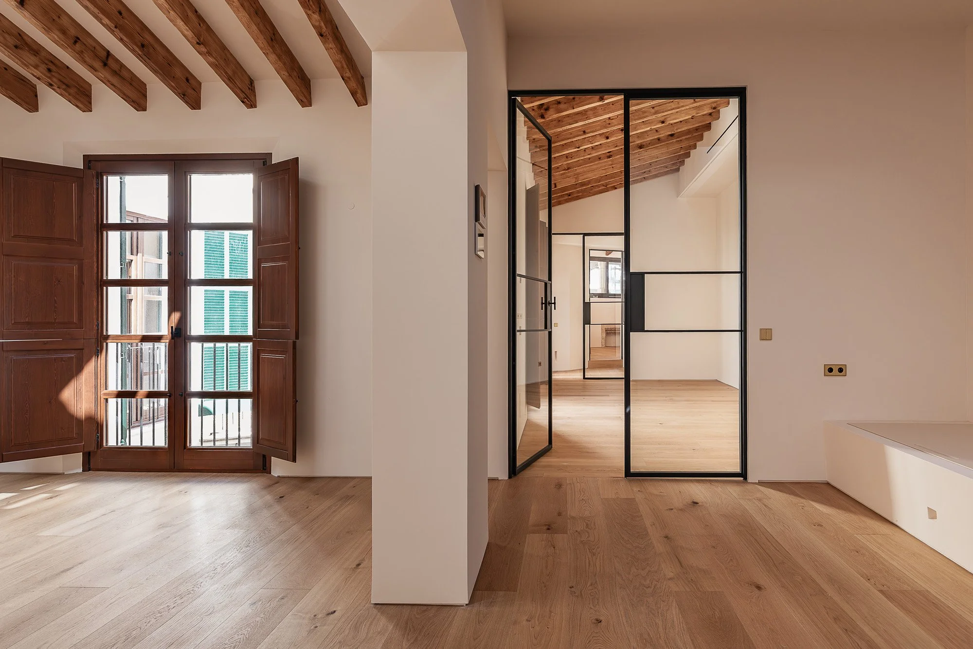 Interior of a modern home with wooden floors, white walls, and a ceiling with exposed wooden beams. There are two sections separated by a black metal-framed glass door, with a window and French doors letting in natural light.