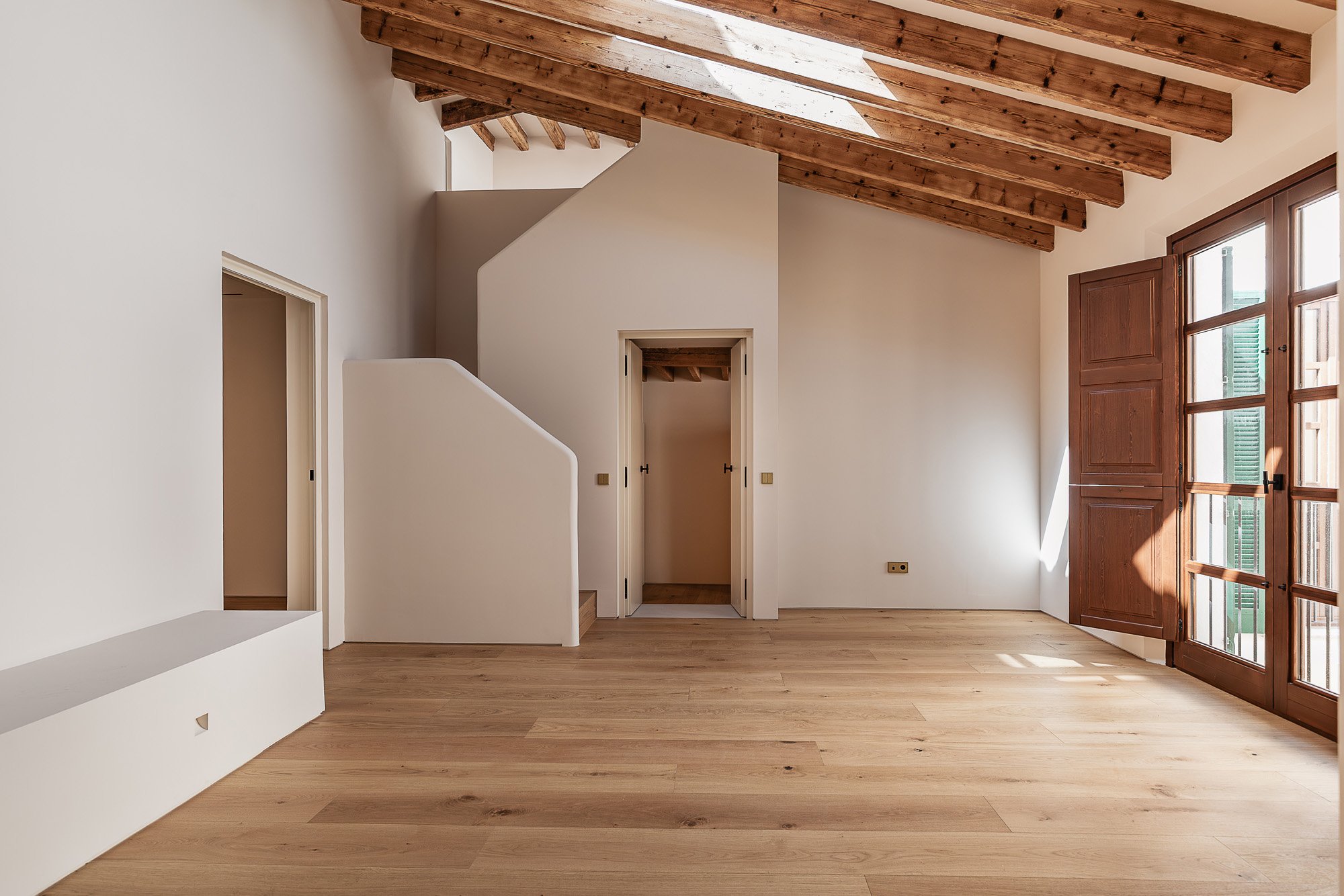 Empty room with wooden floors, white walls, a sloped wooden ceiling, and large glass doors with green shutters.