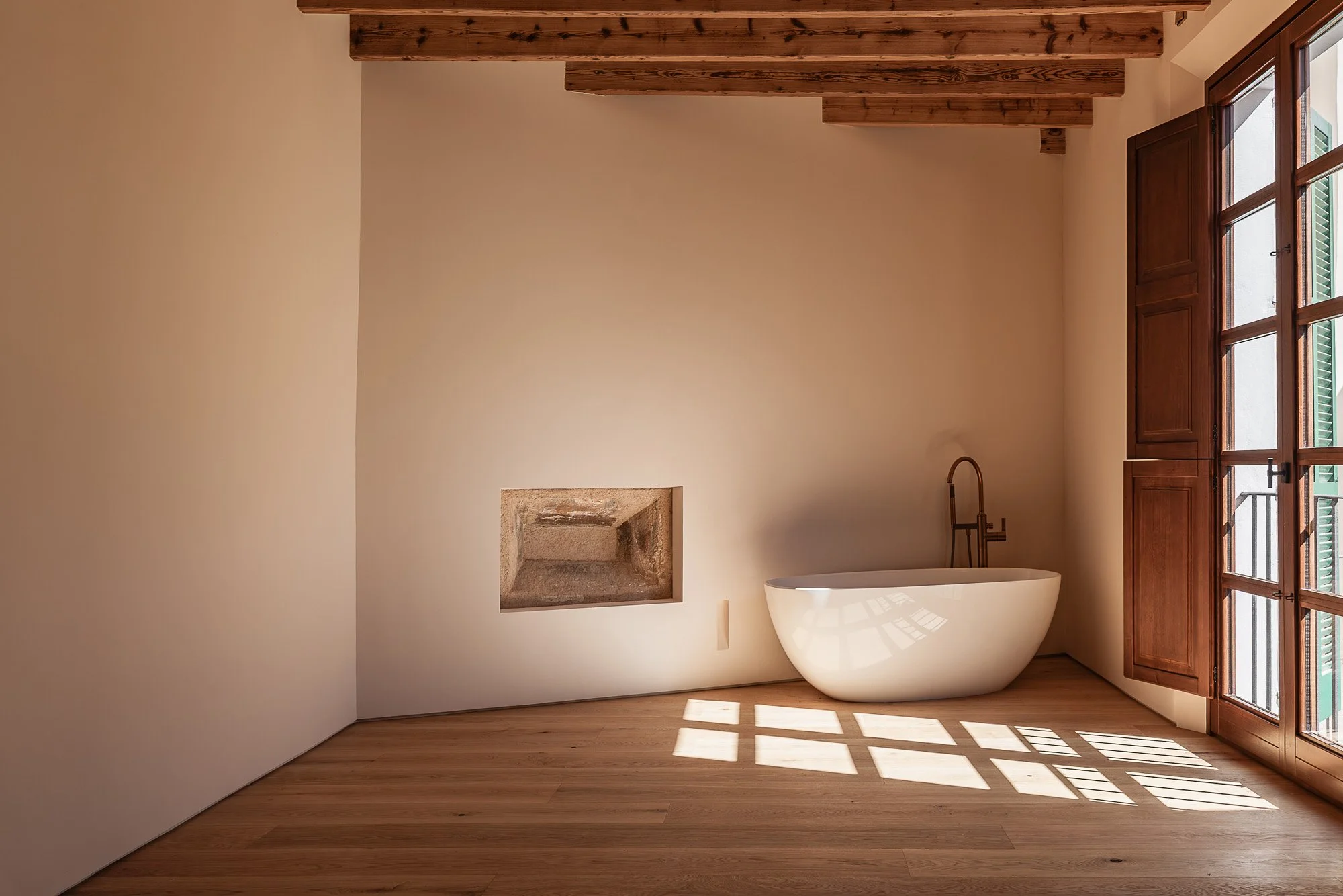 Empty room with a freestanding bathtub, wooden floor, beige walls, and large glass door with green shutters, sunlight casting shadows on the floor, and exposed wooden ceiling beams.