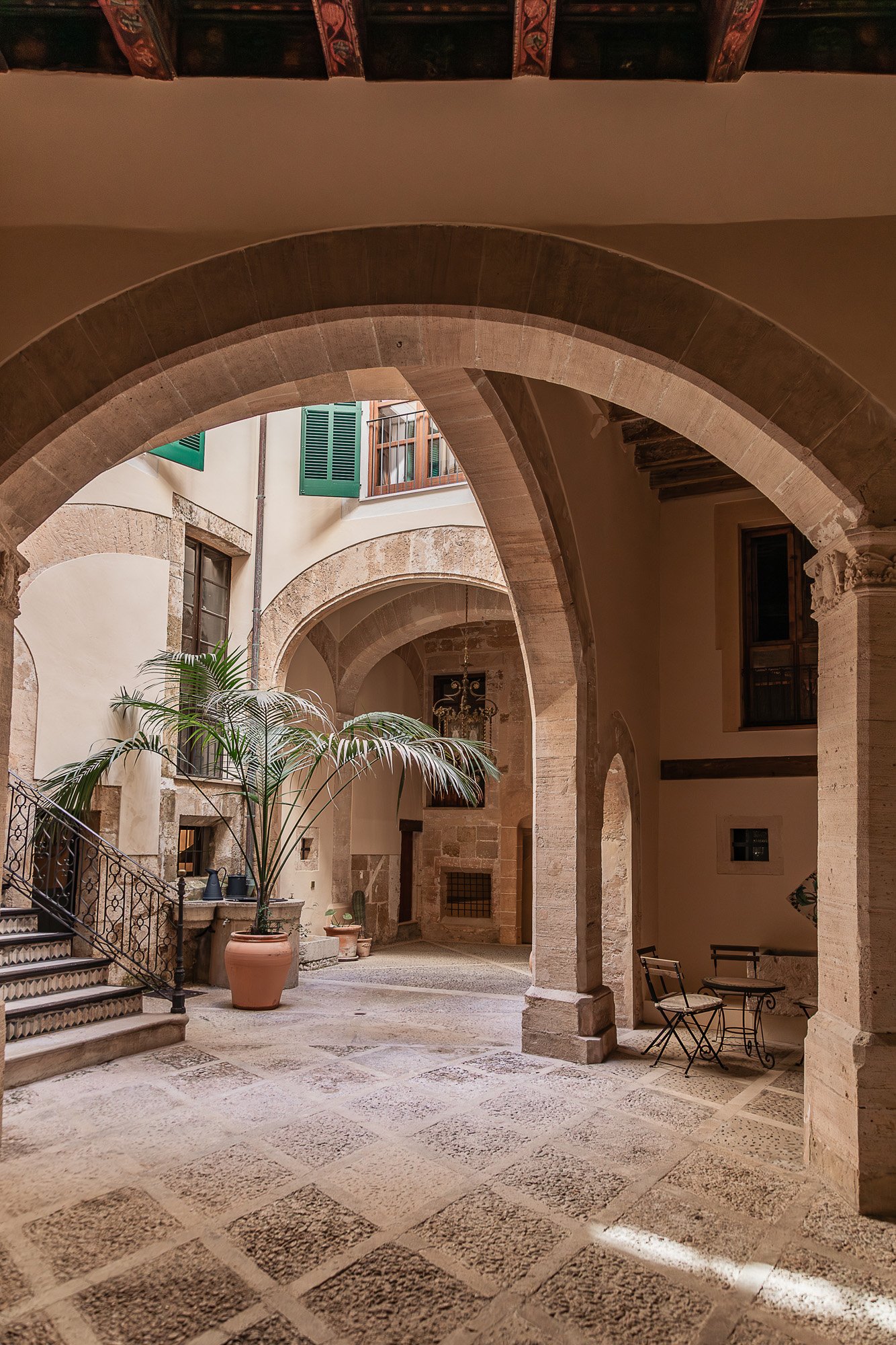 A historic courtyard with stone arches, potted plants including a large palm, and outdoor furniture such as chairs and small tables, featuring a rustic and elegant ambiance.