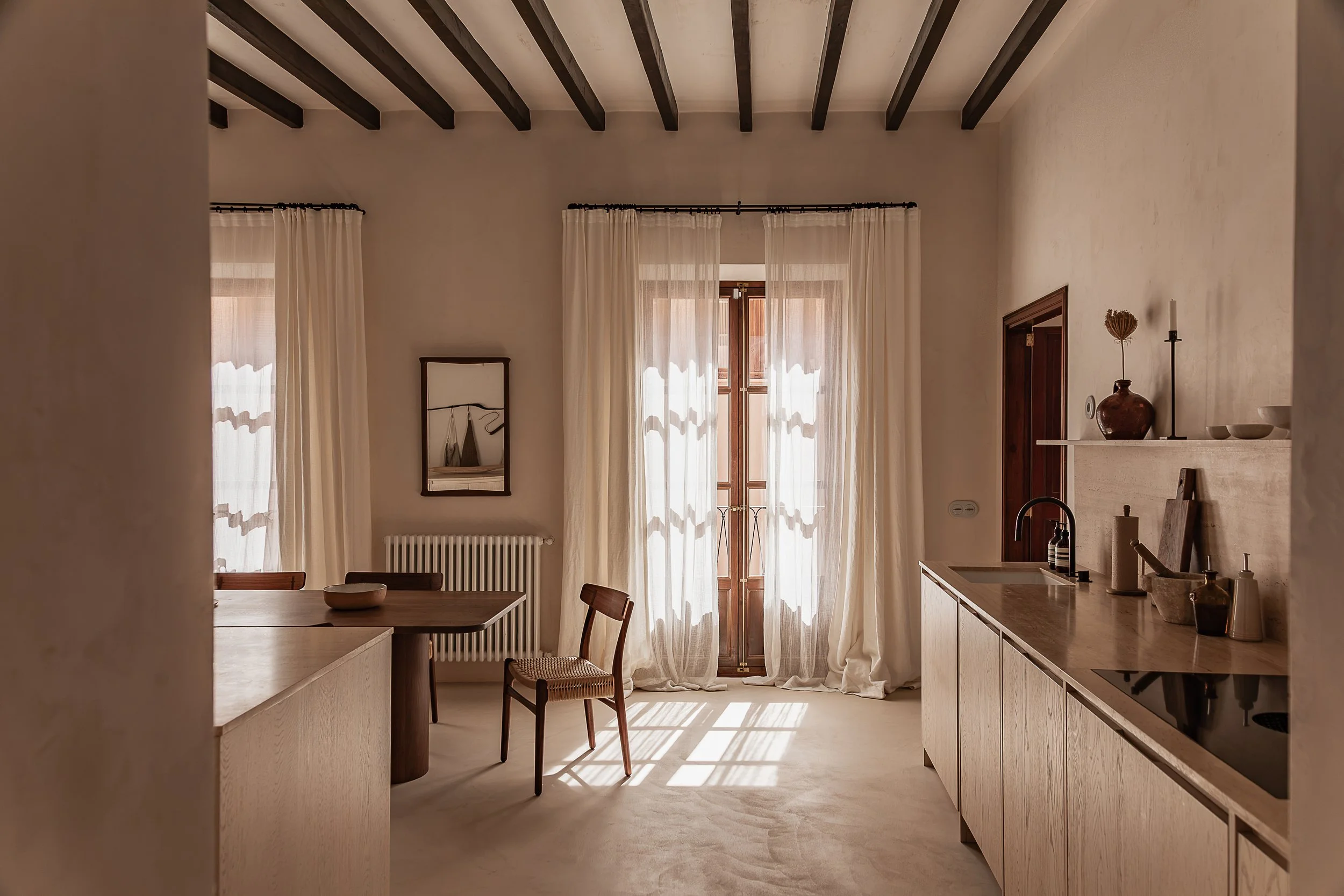 Living room with beige walls, a wooden door, a window with white curtains, a beige armchair, a wooden coffee table with a vase and dried flowers, a white sofa with pillows, a round, striped ottoman, and a textured wall art piece. A large white pendant light hangs from the ceiling with dark wooden beams.