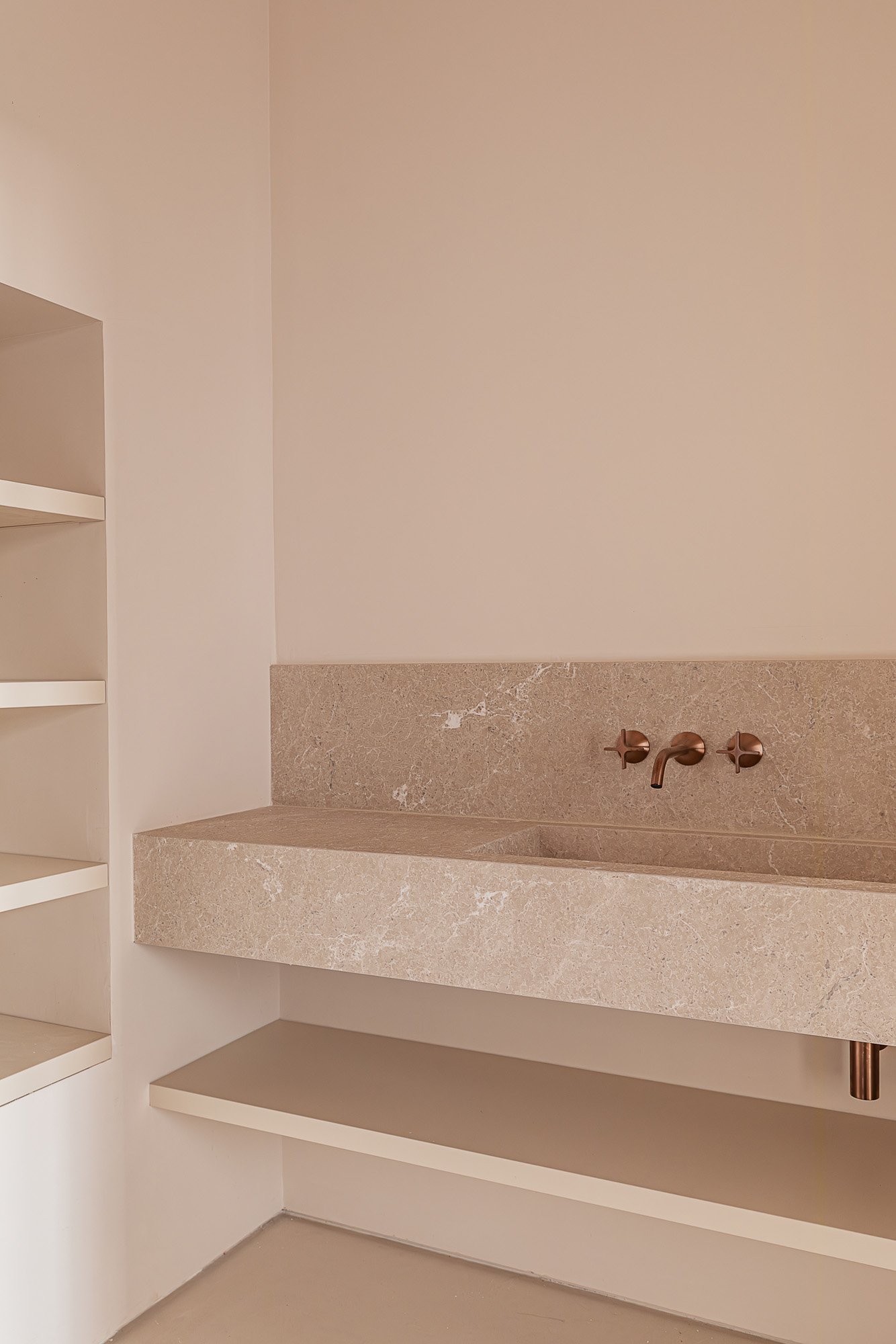A minimalist bathroom with beige marble sink and wall-mounted copper faucet, white wall and open shelves.