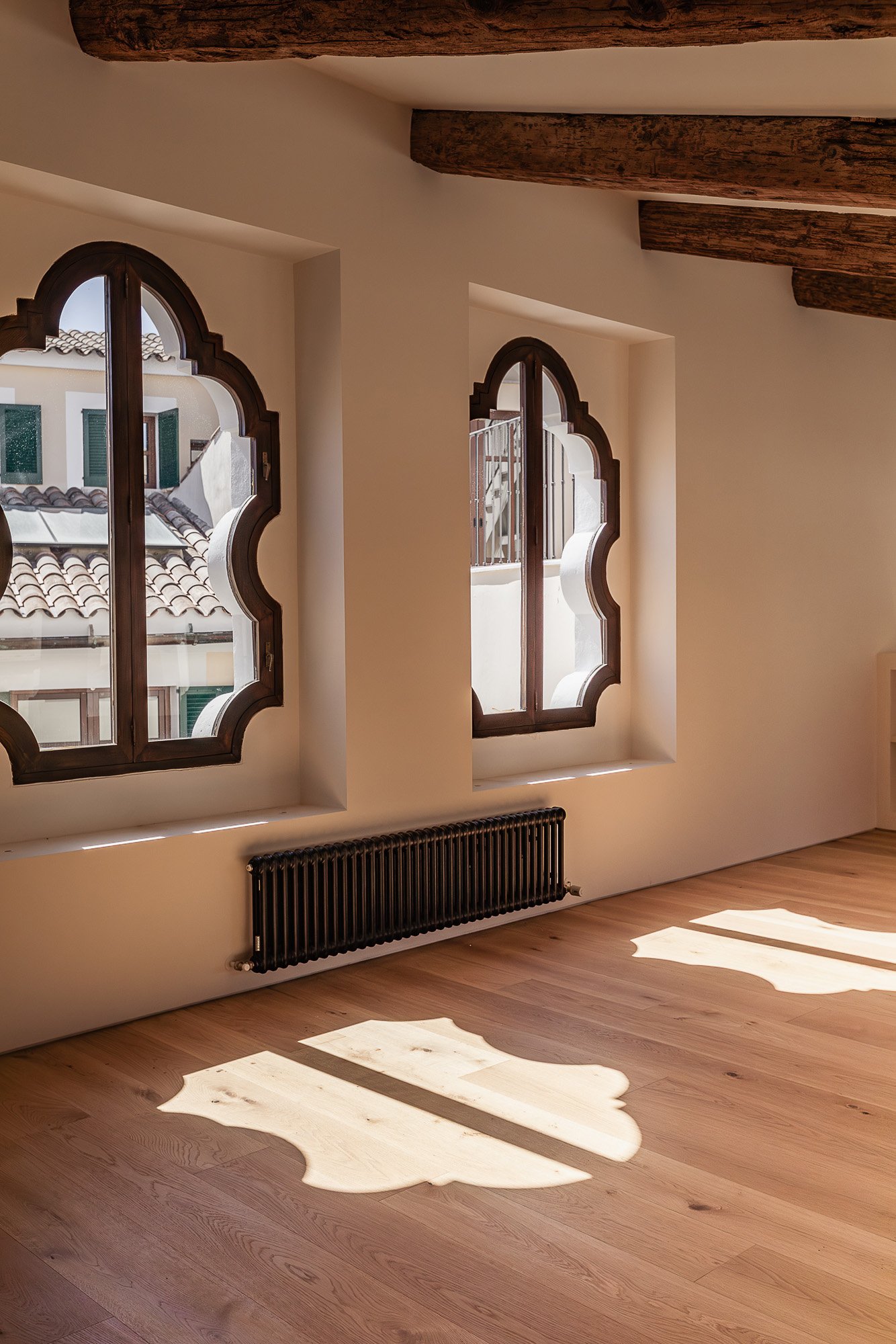 Sunny interior room with two arched windows, wooden beams on ceiling, hardwood floor, and a black radiator beneath the windows, with sunlight casting decorative shadows on the floor.