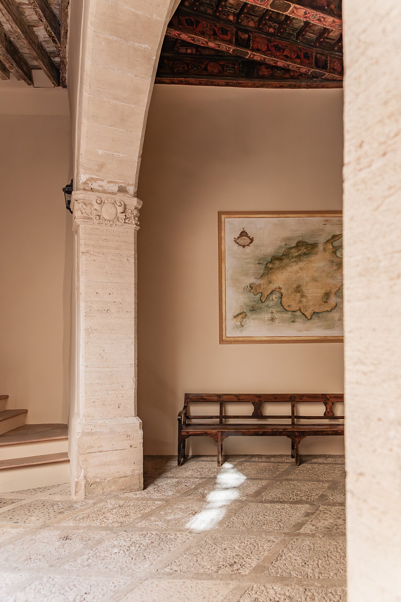 Interior corner of a historic stone building with a beige wall, a wooden bench, and a framed antique map of an island on the wall. Sunlight creates bright patches on the stone floor.
