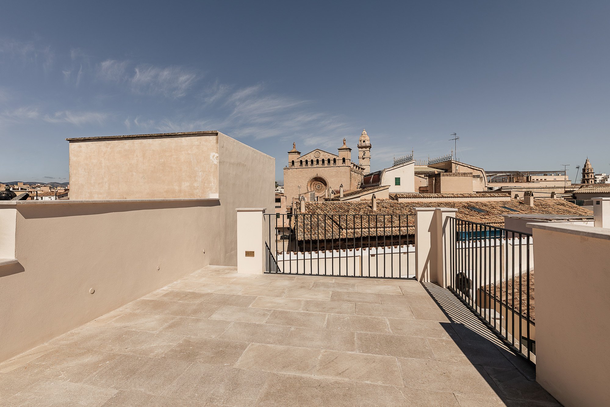 View of rooftop terrace with beige tiled floor and black metal railing overlooking historic European city skyline with church and bell towers under blue sky.