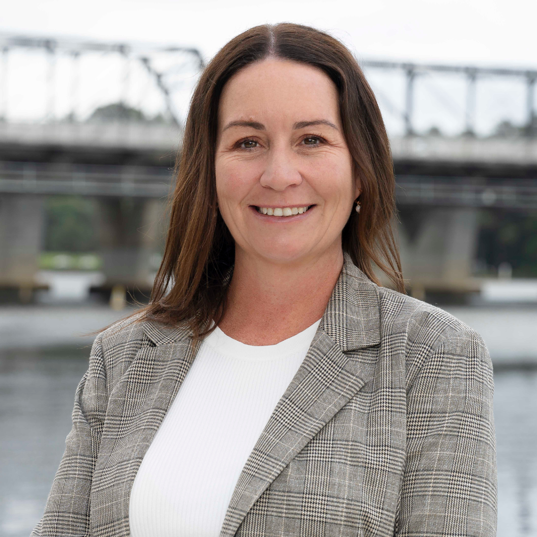 A woman standing outdoors in front of green foliage, wearing a plaid blazer and black top, smiling slightly, with her hair pulled back and hoop earrings.