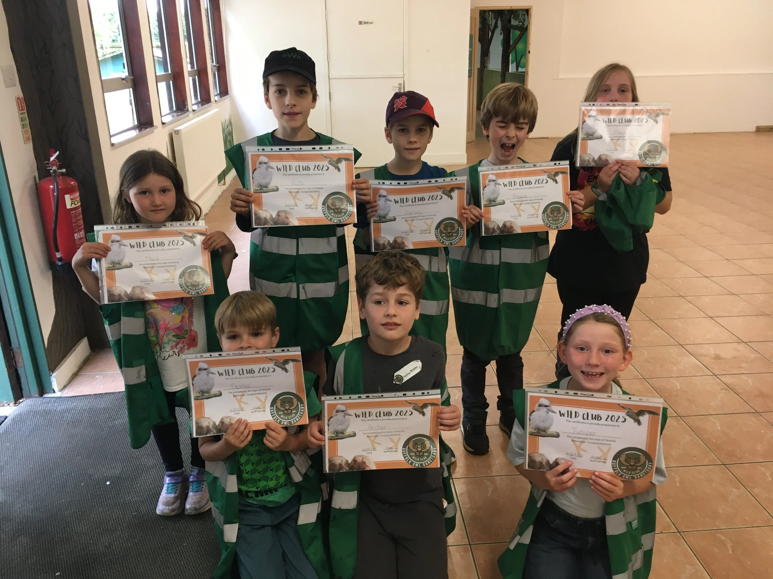 Group of children holding certificates for a wild club 2023 event, standing indoors on tiled floor.
