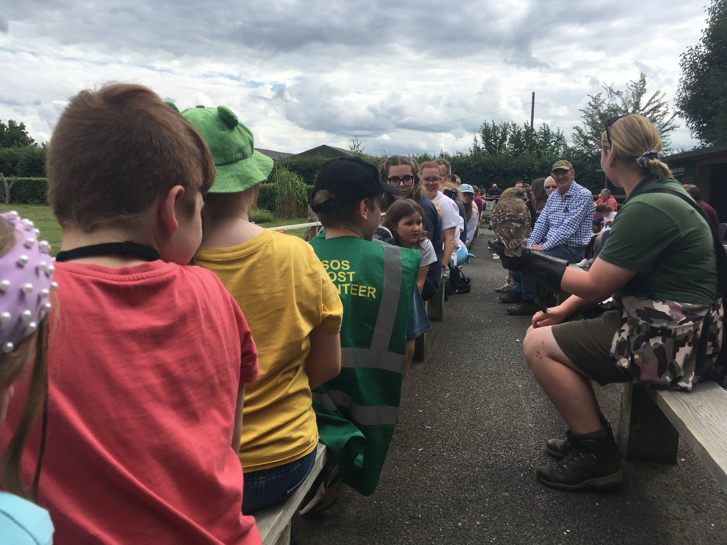 A group of children and adults sitting on benches outdoors listening to a woman in uniform holding a bird of prey during a wildlife presentation.