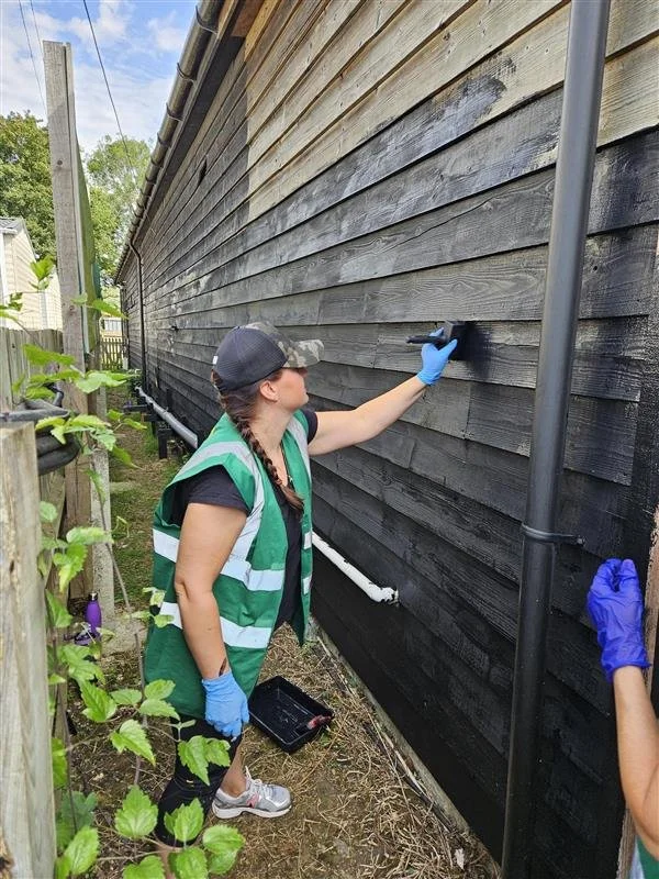 Person painting the exterior of a black wooden house with a paintbrush, wearing a green safety vest, blue gloves, and a cap, standing outdoors during daytime.