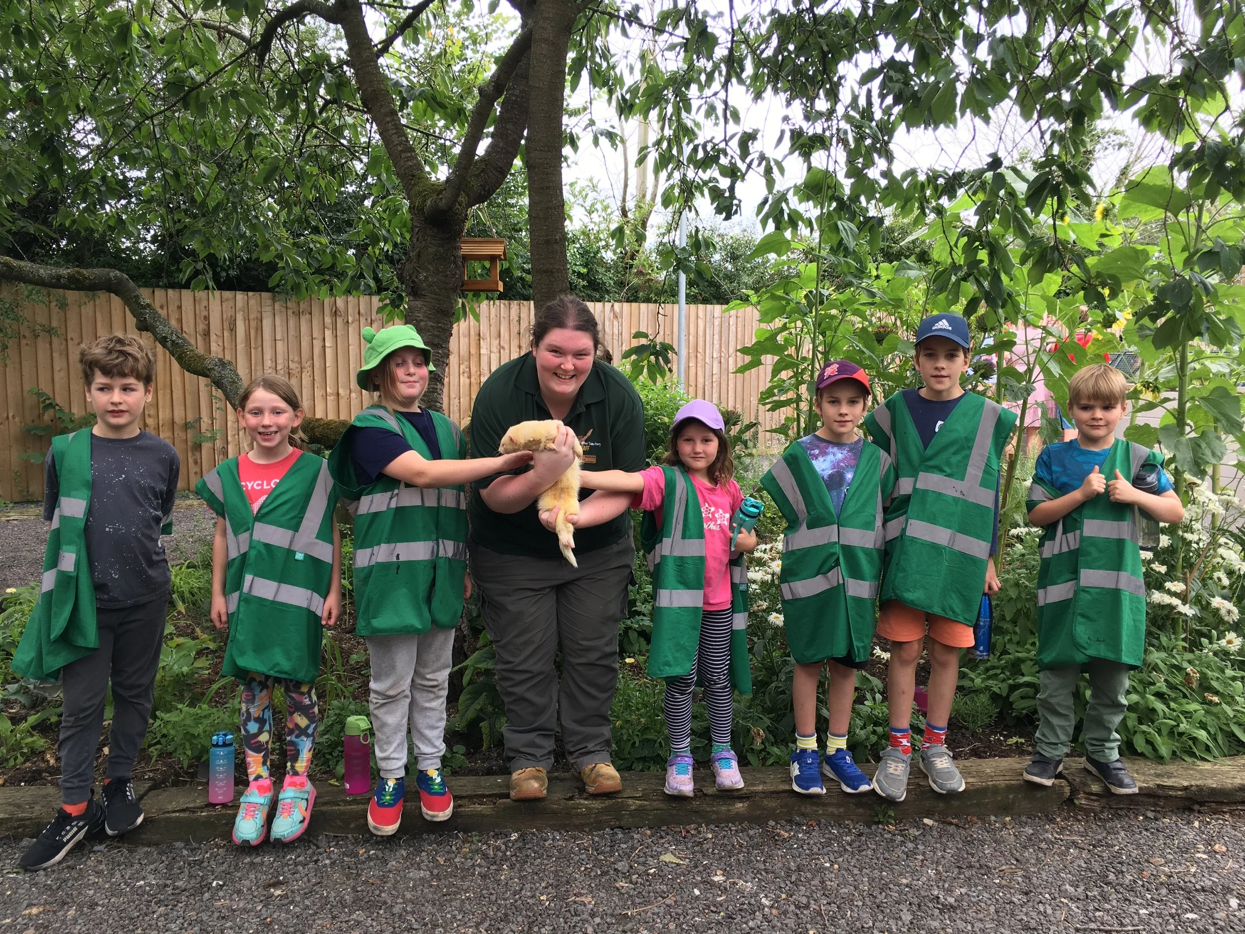 Group of young children and a woman outdoors in a garden, wearing green safety vests. The woman is holding a yellow duckling, and children are reaching out to touch it. The background includes trees, bushes, and a wooden fence.