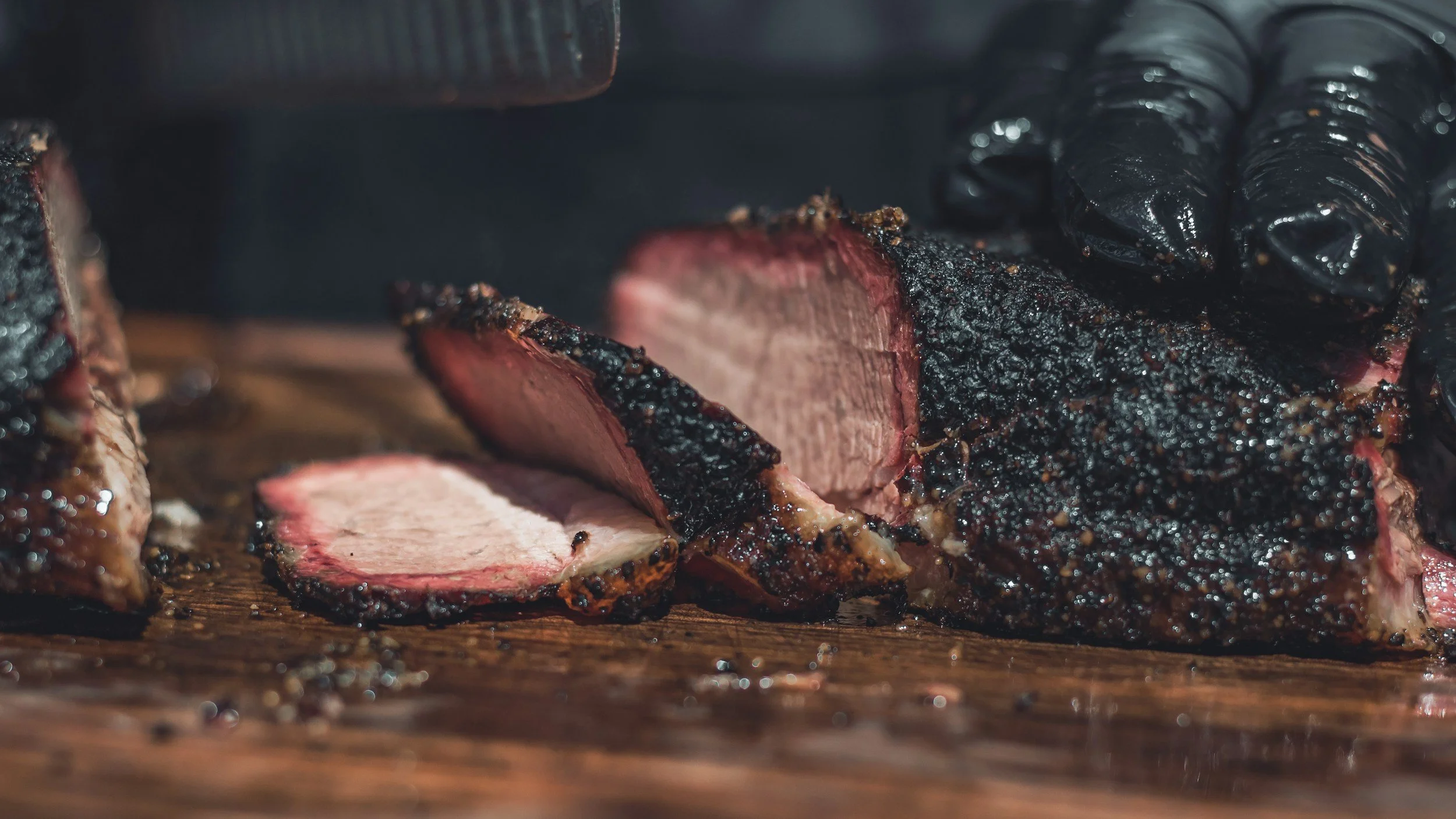Close-up of a smoked beef brisket with a dark, charred crust, sliced on a wooden surface, with black barbecue sauce nearby.