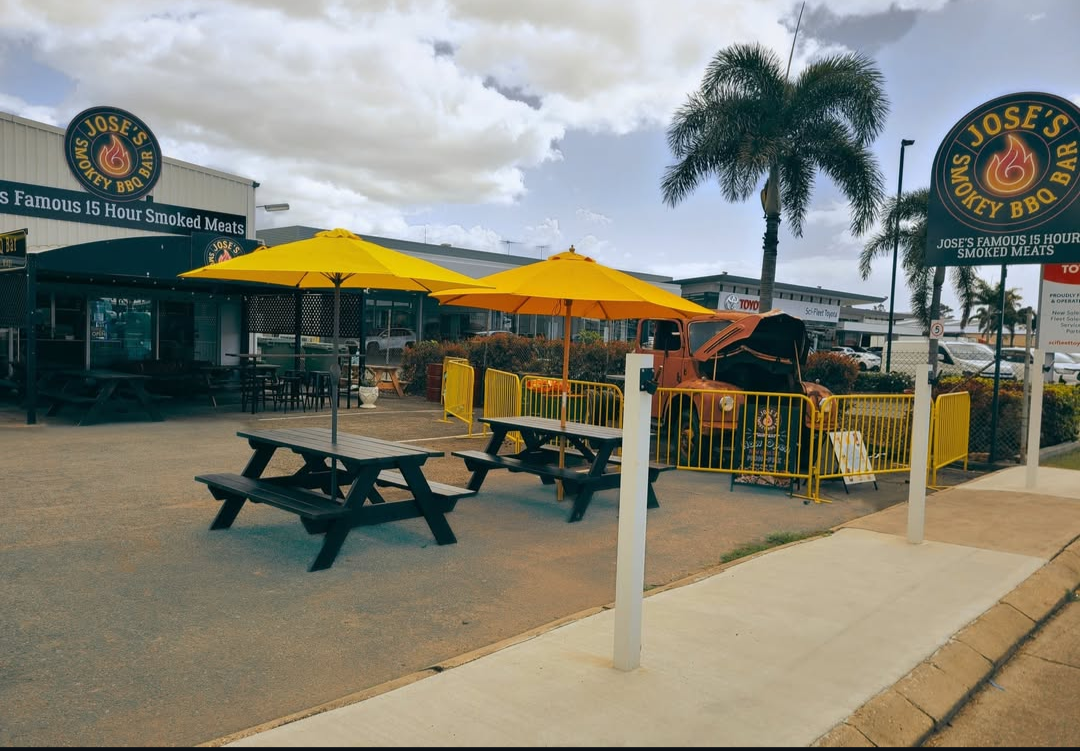 Outdoor seating area in front of a BBQ restaurant with yellow umbrellas, picnic tables, palm trees, and a vintage orange car displayed behind a yellow barrier.