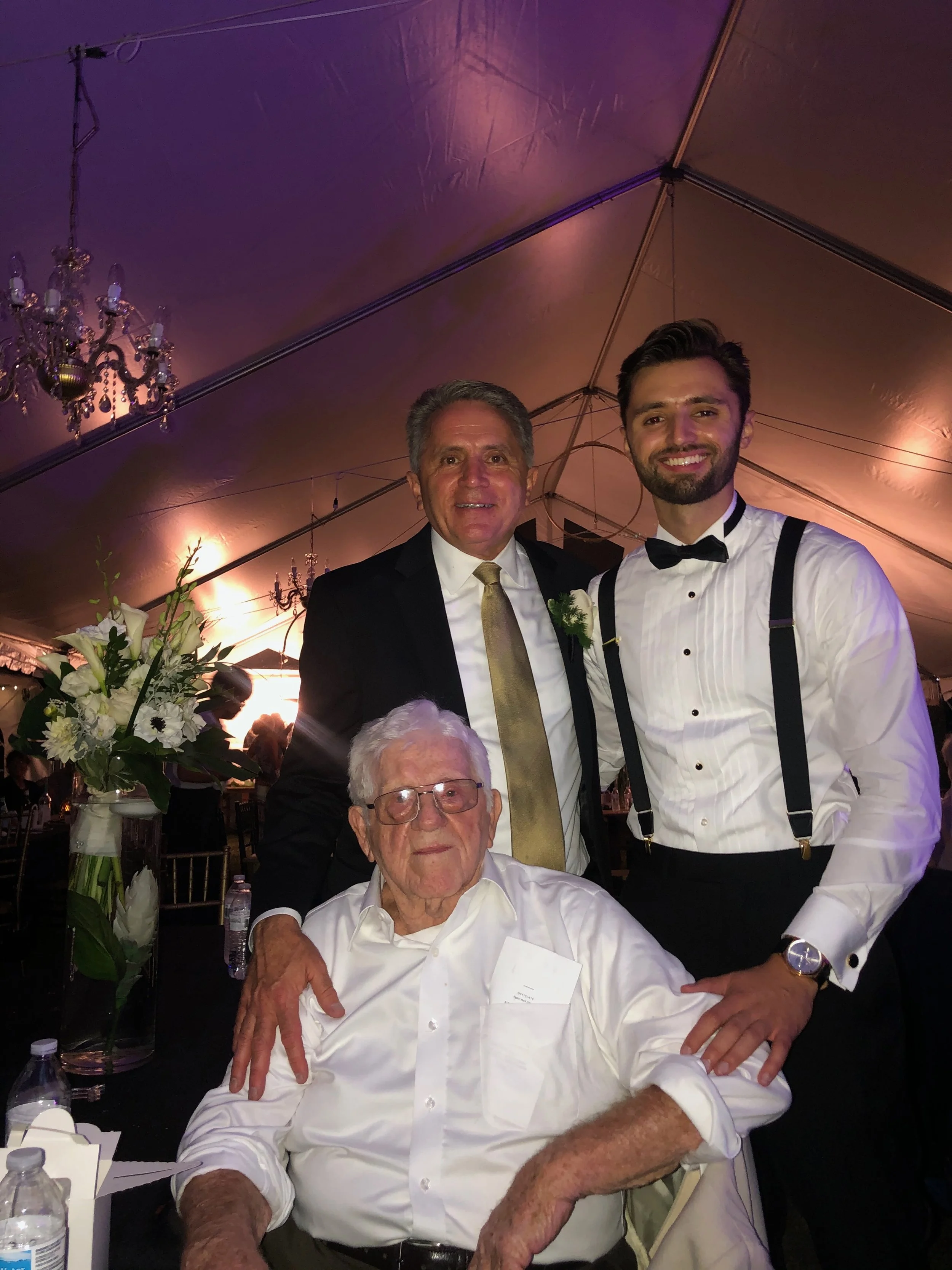 Three men dressed in formal attire at a wedding reception in a decorated tent. One man is seated and wearing glasses, while the two others are standing behind him. The standing man on the right is wearing a tuxedo with suspenders and a bow tie. The man on the left is wearing a black suit with a gold tie, and the seated man has a white shirt.
