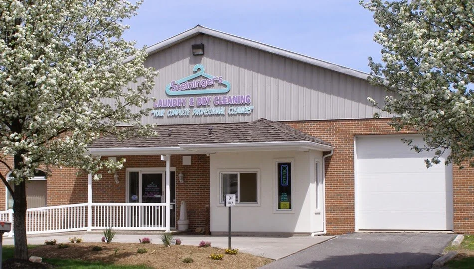 Exterior of a laundry and dry cleaning business called 'Steininger's' with a brick and gray siding building, a small front porch, and trees with white blossoms.