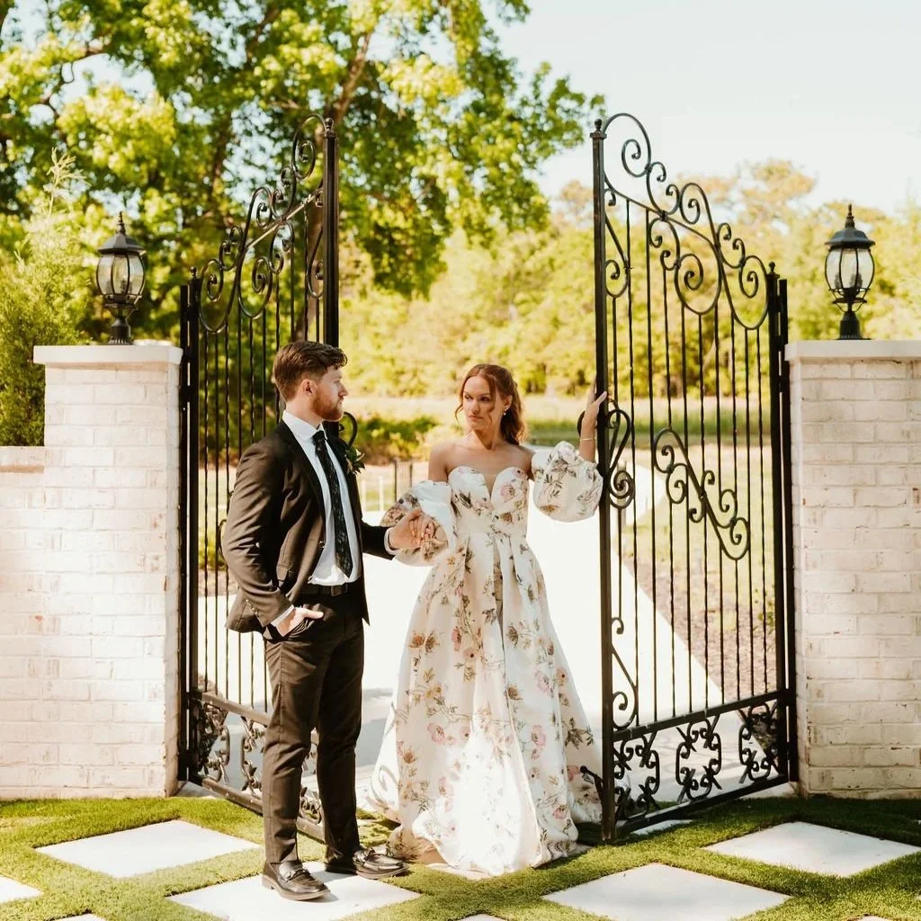 A bride and groom standing at an ornate wrought iron gate outdoors on a sunny day, surrounded by greenery.