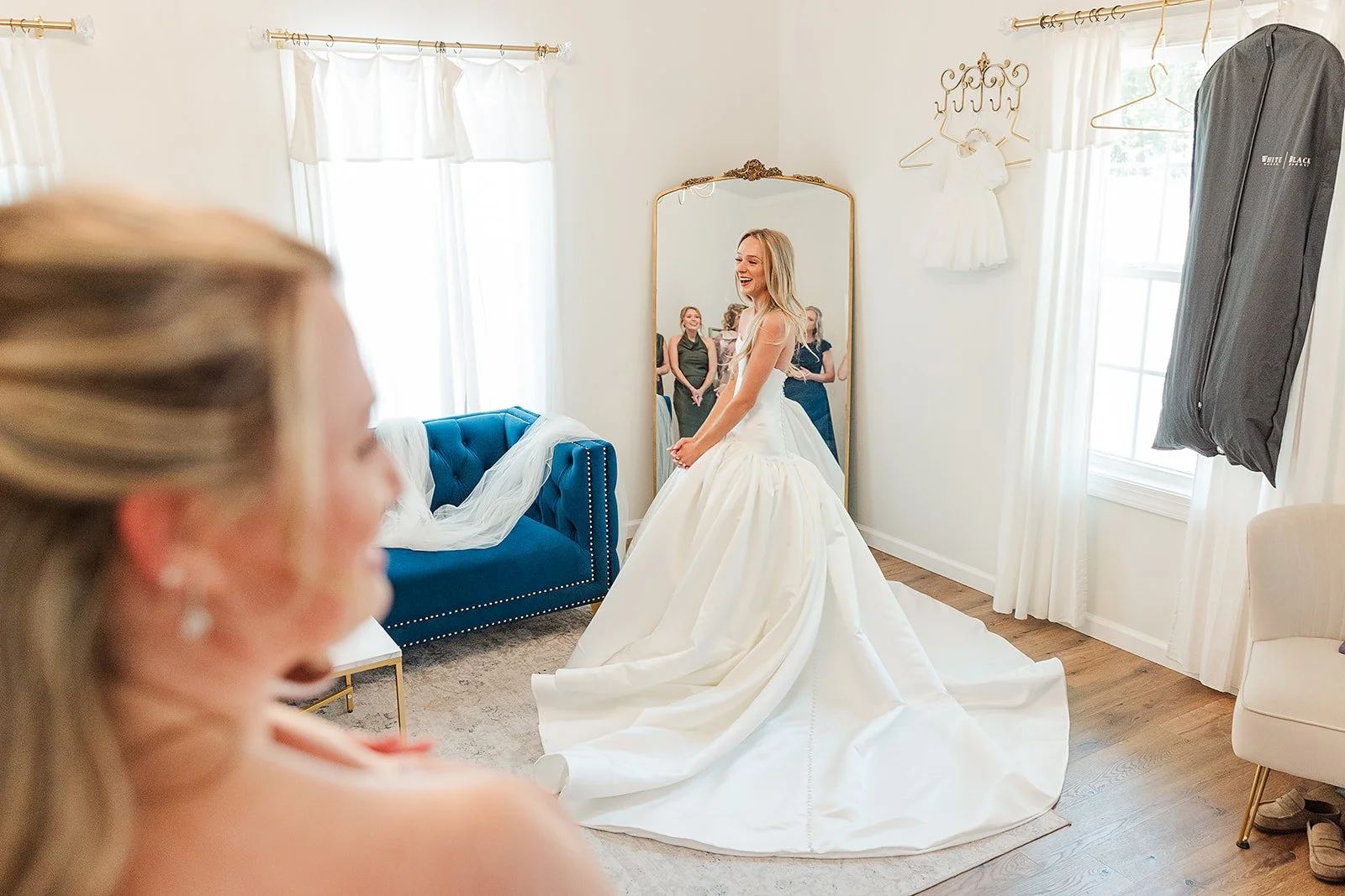 A bride in a white wedding dress laughs in front of a mirror while her bridesmaids watch in a bright, well-lit room with white curtains and modern furniture.