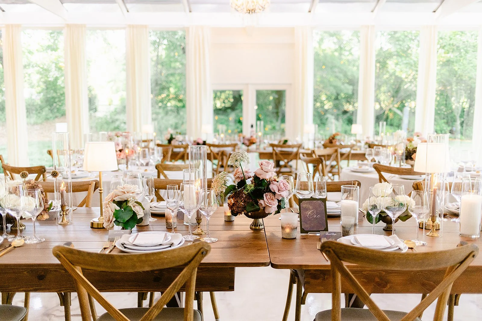 Wedding reception table setting with floral centerpieces, candles, wine glasses, and place settings in a bright sunroom with large windows and greenery outside.