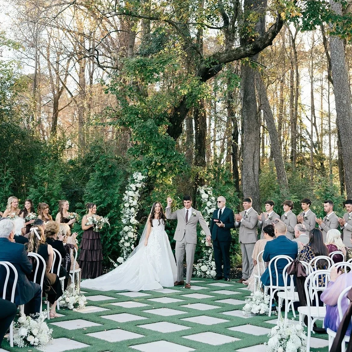 Wedding ceremony outdoors in a forest setting, with a bride and groom standing beneath a floral arch, surrounded by bridesmaids and groomsmen, guests seated on white chairs decorated with white flowers.