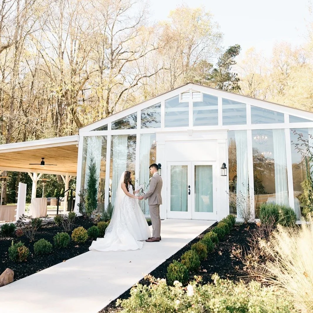 A bride and groom holding hands and exchanging vows outside a white glass chapel surrounded by landscaped bushes and trees.