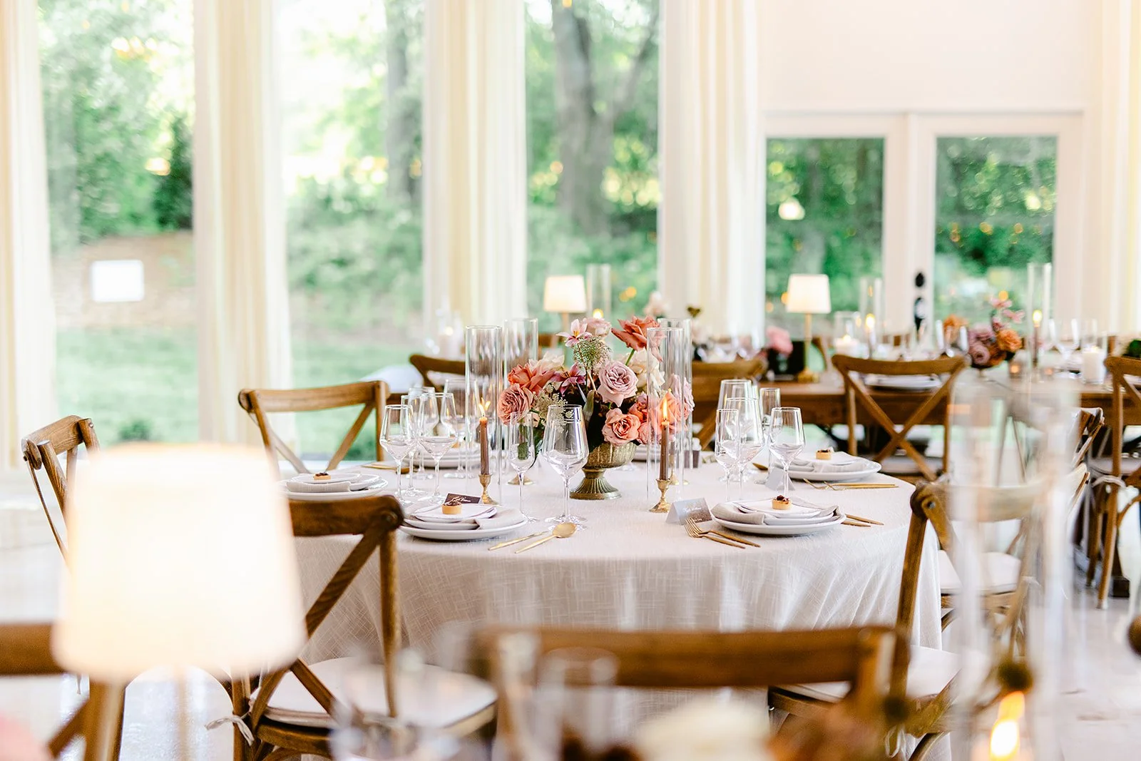 A dining table set for a formal event, decorated with pink and white flowers in a vase, surrounded by wooden chairs, with tableware including wine glasses, candles, and napkins, inside a bright room with large windows showing a green outdoor scene.
