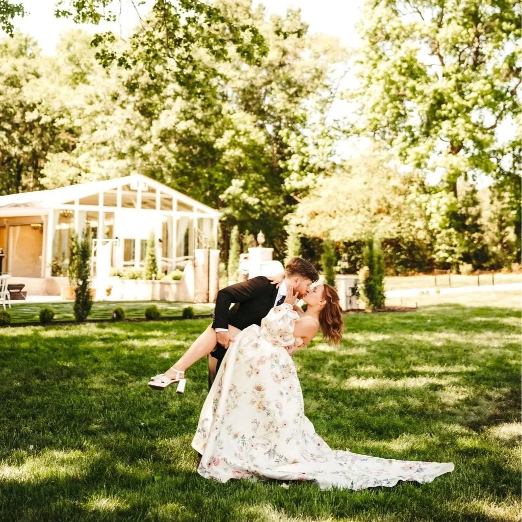 A bride and groom sharing a kiss outdoors on a grassy lawn, with the groom dipping the bride. The bride wears a floral wedding dress, and the groom is in a black suit. There are trees and a glass pavilion in the background.