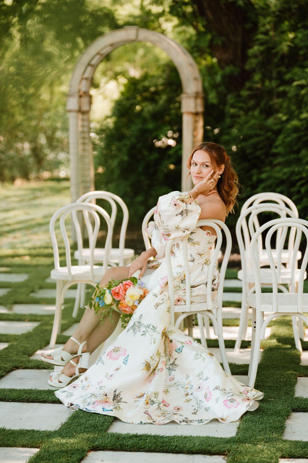 A woman in a floral dress sitting on a white chair outdoors, holding a bouquet of flowers, with greenery and a stone archway in the background.