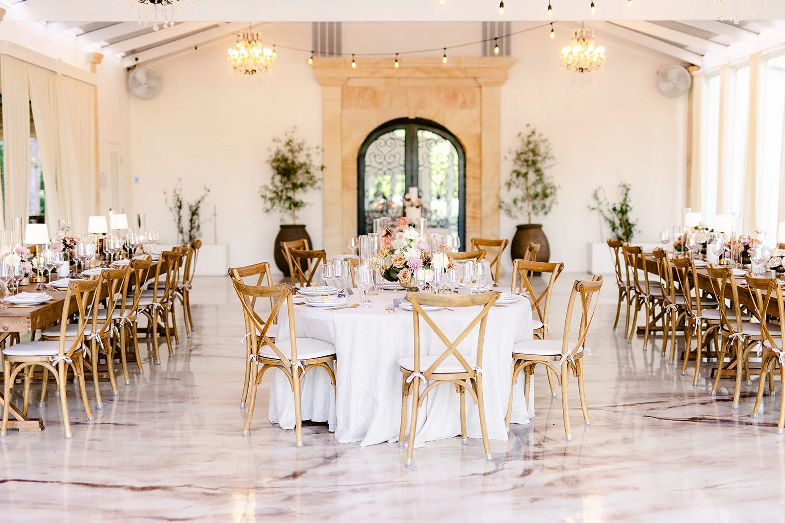 A banquet hall decorated for a wedding reception with a large round table in the center with pink and white floral arrangements, surrounded by wooden chairs with white cushions, and long rectangular tables along the sides with similar floral centerpieces. The room features white walls, large windows, chandeliers, and potted plants.
