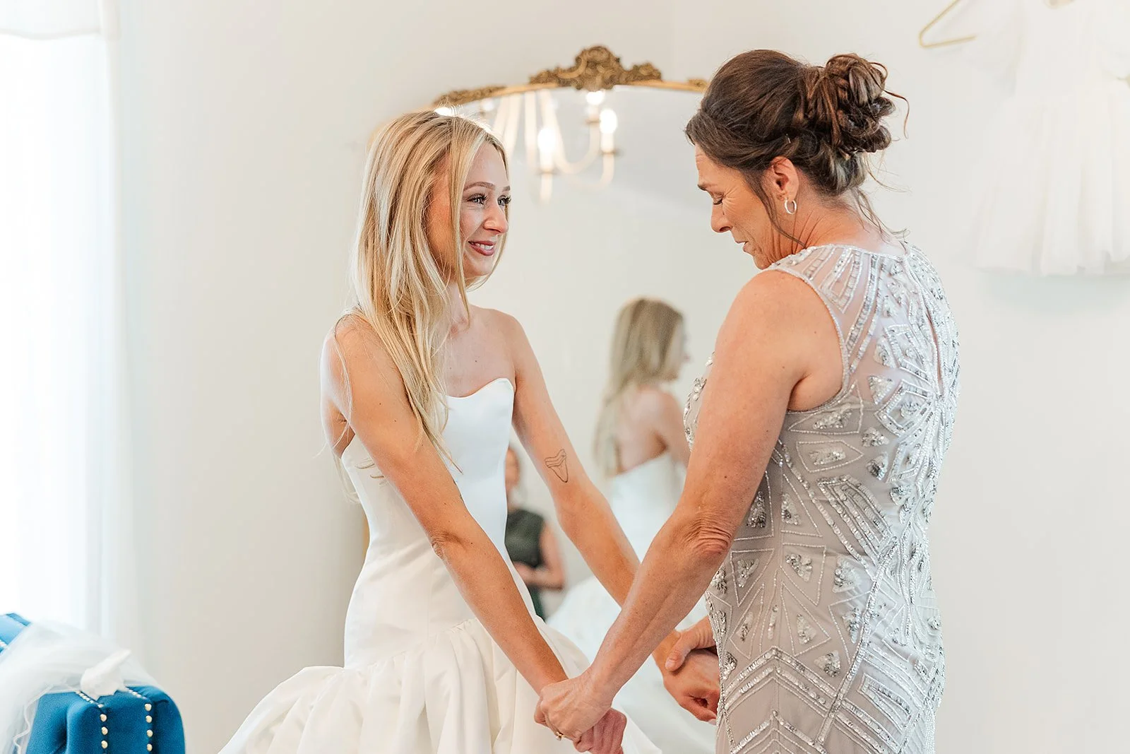 A bride holding hands with an older woman, possibly her mother, on her wedding day. They are inside a room with a mirror and a chandelier reflected in the background, both dressed in elegant wedding attire.