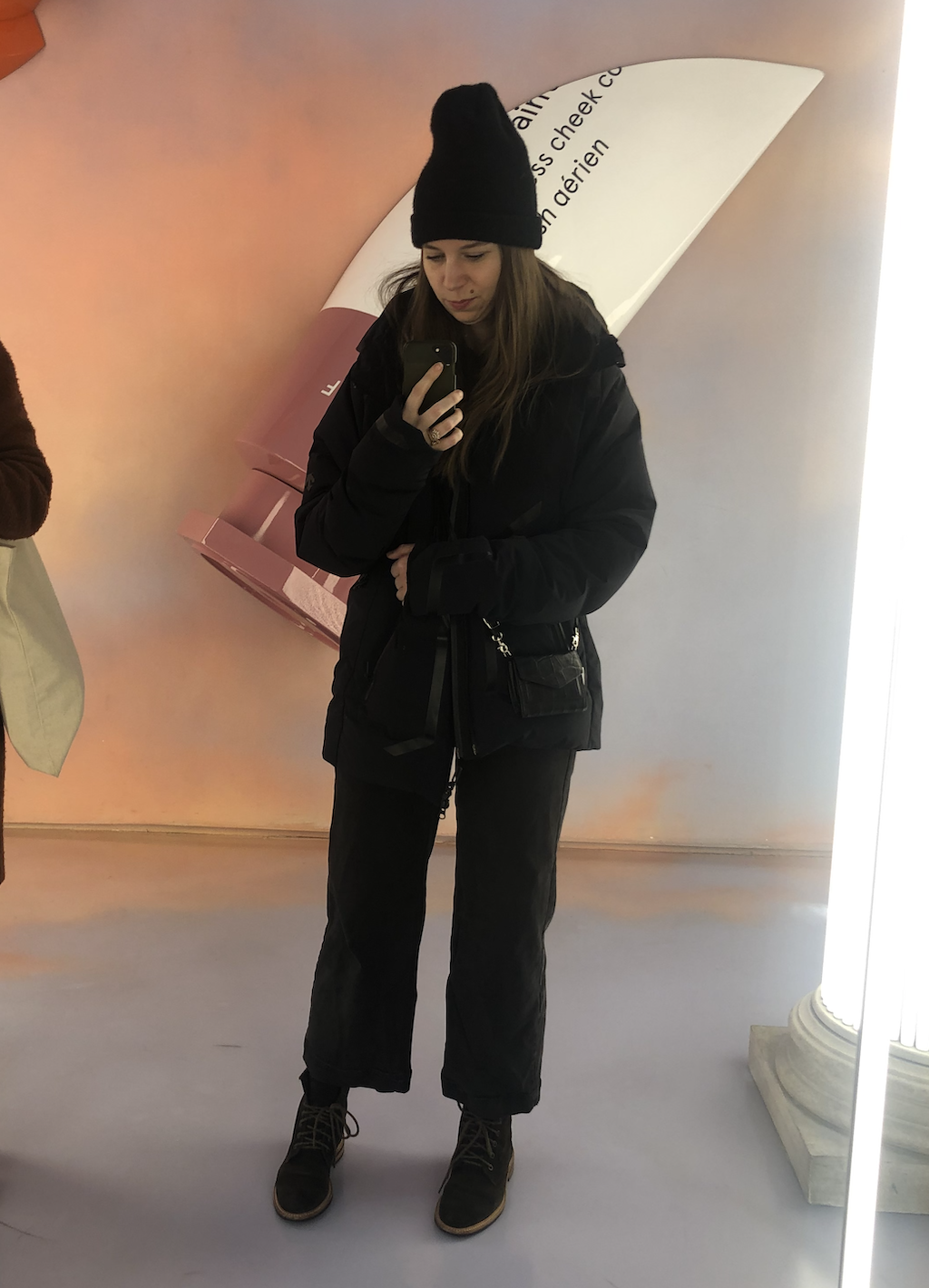 A woman with long brown hair taking a selfie in an elevator mirror. She is wearing a black beanie, black jacket, black pants, and boots. Behind her is a colorful wall with a large, abstract, boat-shaped graphic and text.