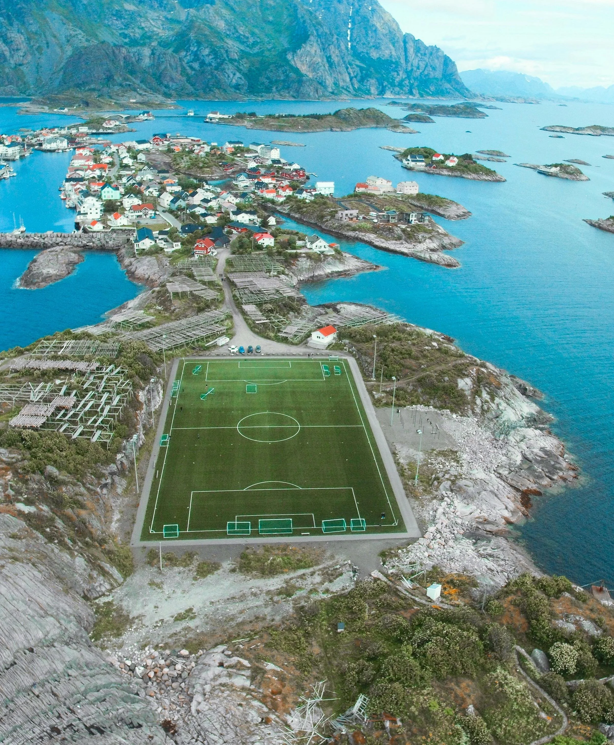 A football field built on a rocky hillside overlooking a coastal village with colorful houses and water surrounding the area.