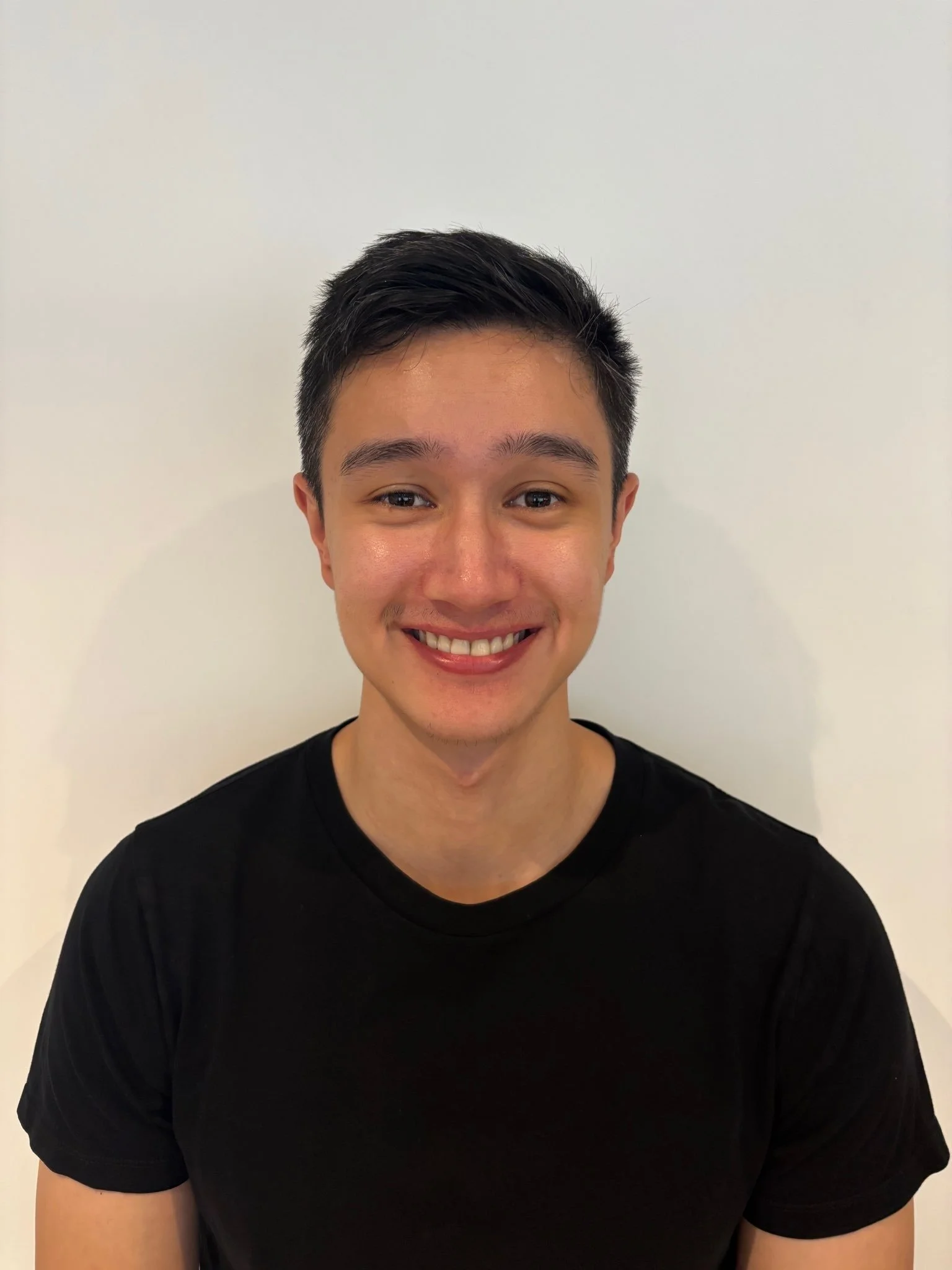 Young man smiling, wearing a black t-shirt, against a plain white wall.