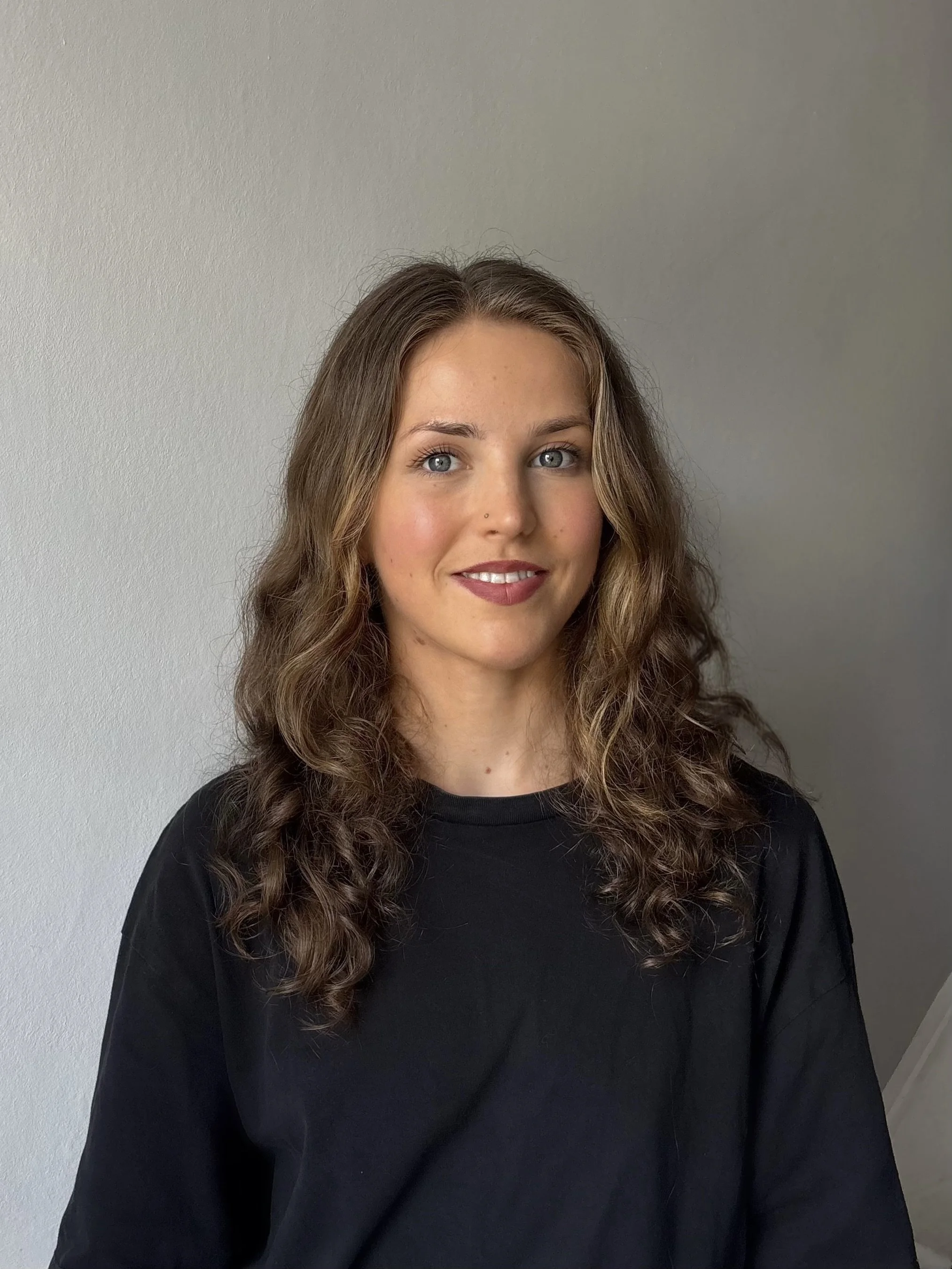 Portrait of a young woman with wavy brown hair, blue eyes, wearing a black top, smiling, against a plain wall background.