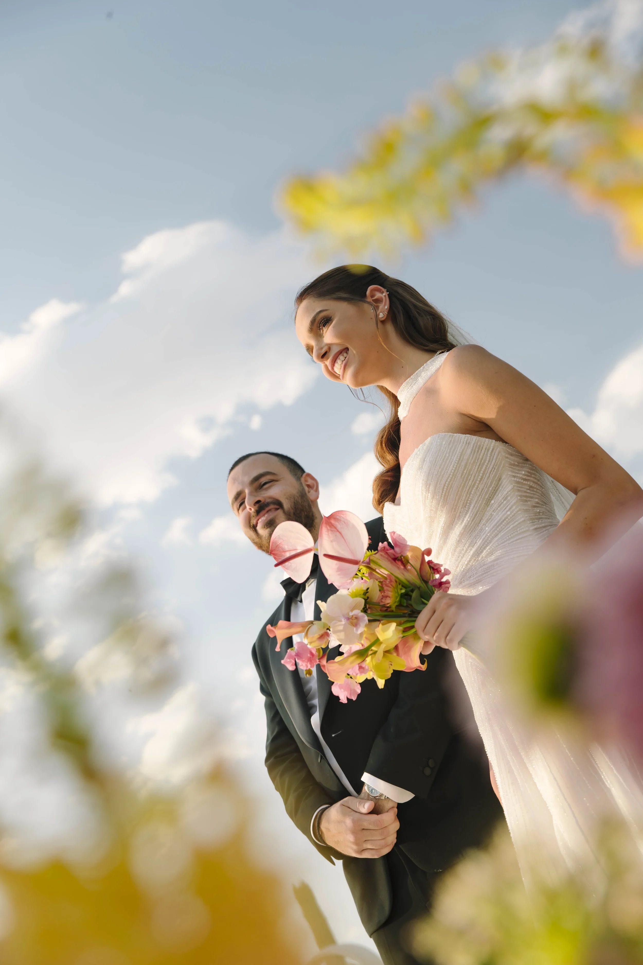 Pareja de recién casados sonriendo, la novia sosteniendo un ramo de flores, en un día soleado con cielo azul y algunas nubes.