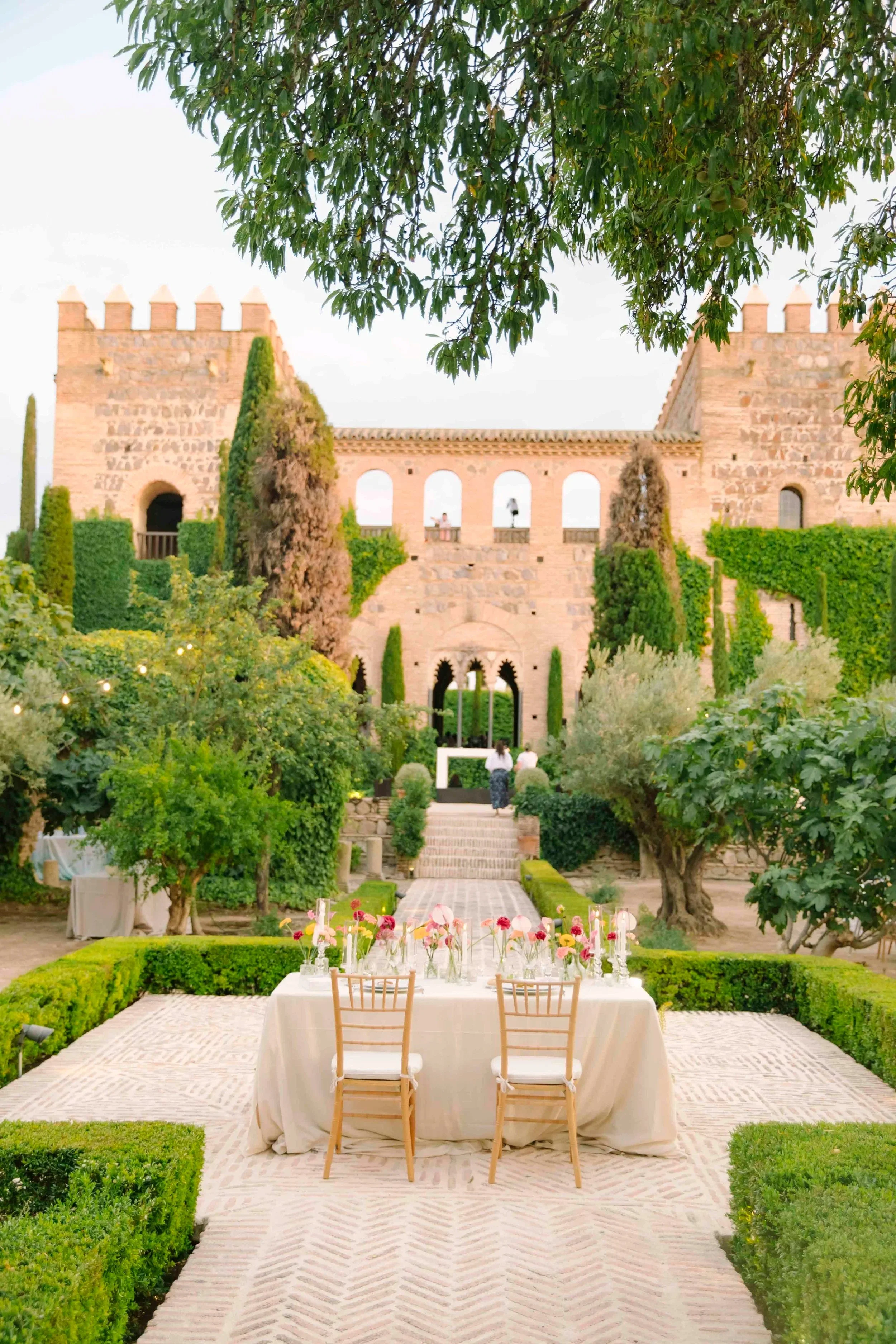 Castillo de Galiana en Toledo Escenario al aire libre con mesa y sillas, decorado con flores, en un jardín cercano a un castillo de piedra con torres y muros, rodeado de árboles y arbustos verdes.