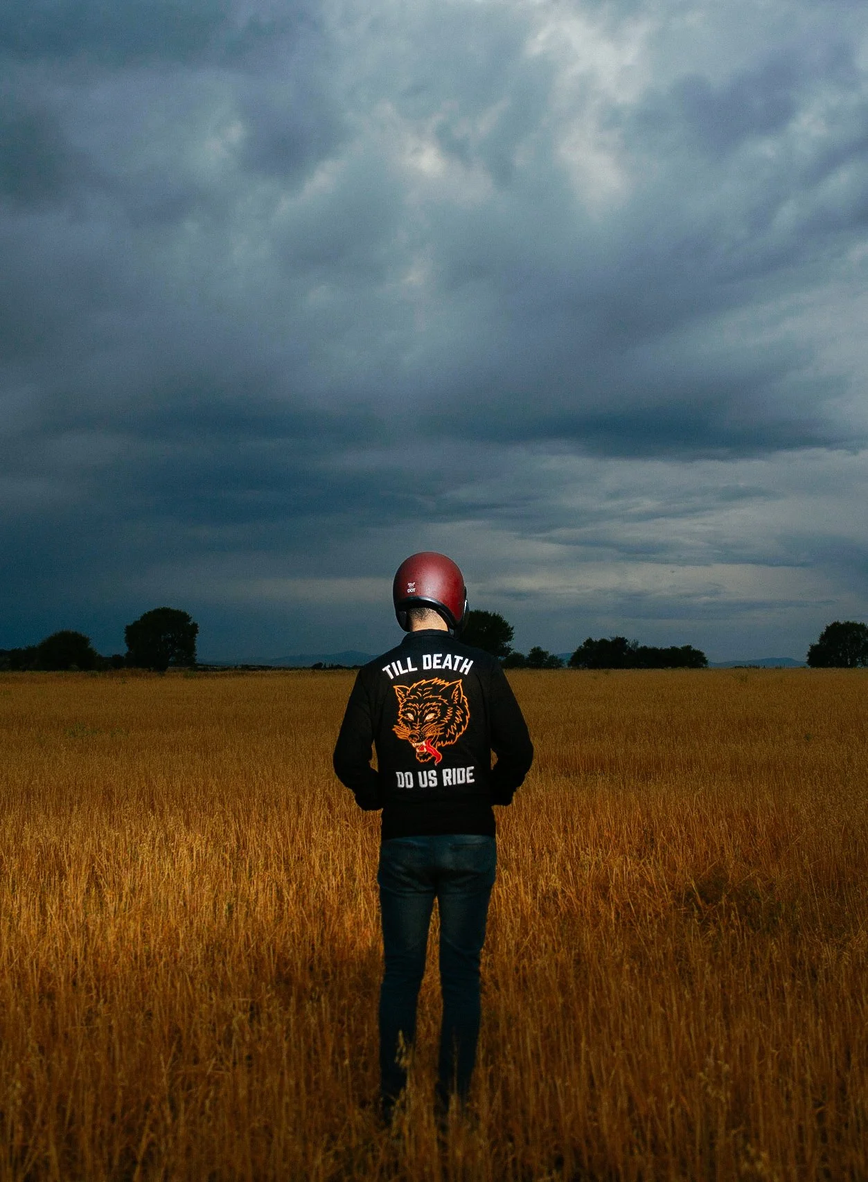 Persona con casco rojo y chamarra negra en campo de hierba dorada, cielo nublado.