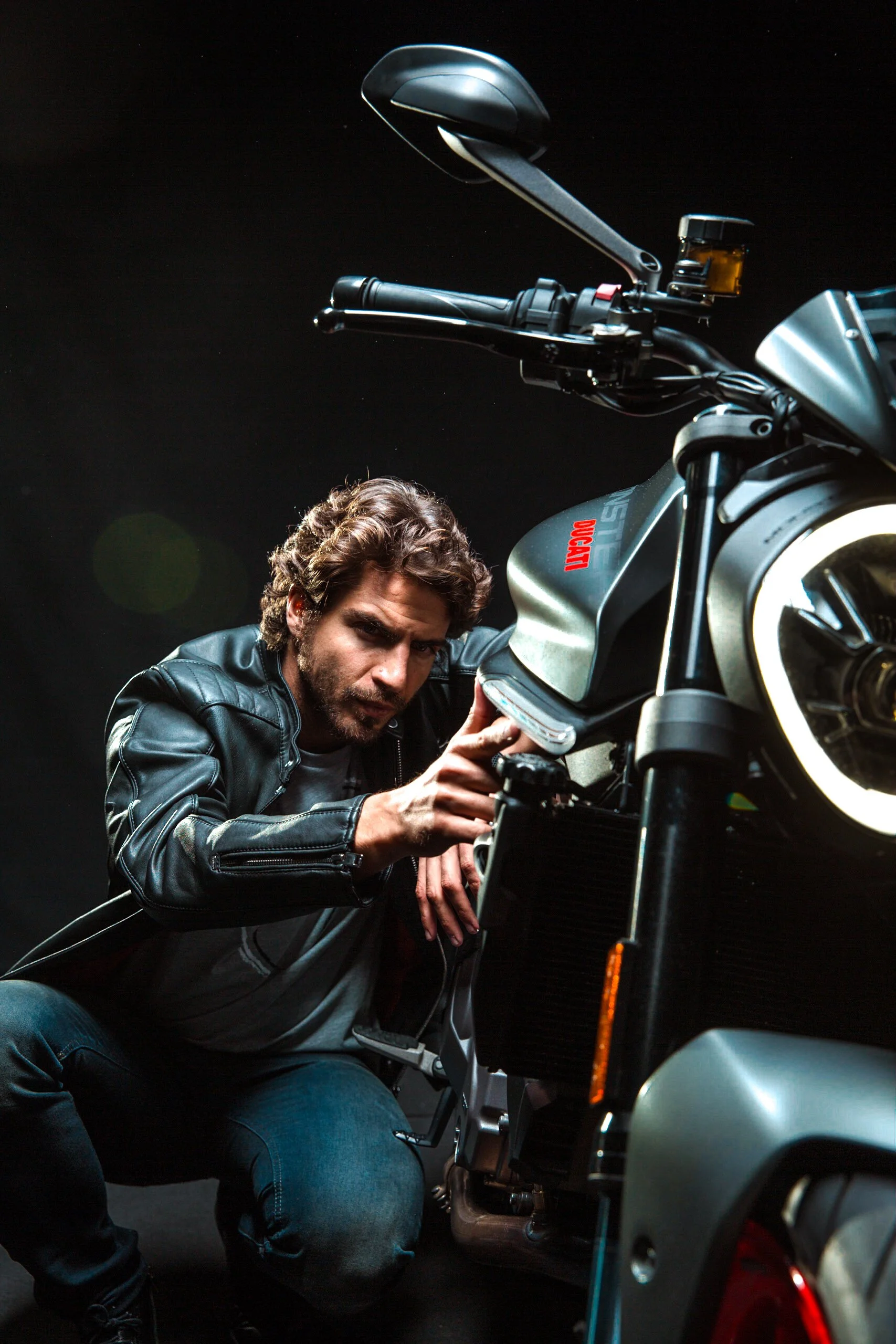 Hombre con cabello castaño y barba, usando chaqueta de cuero, inspeccionando una motocicleta moderna en un fondo negro.