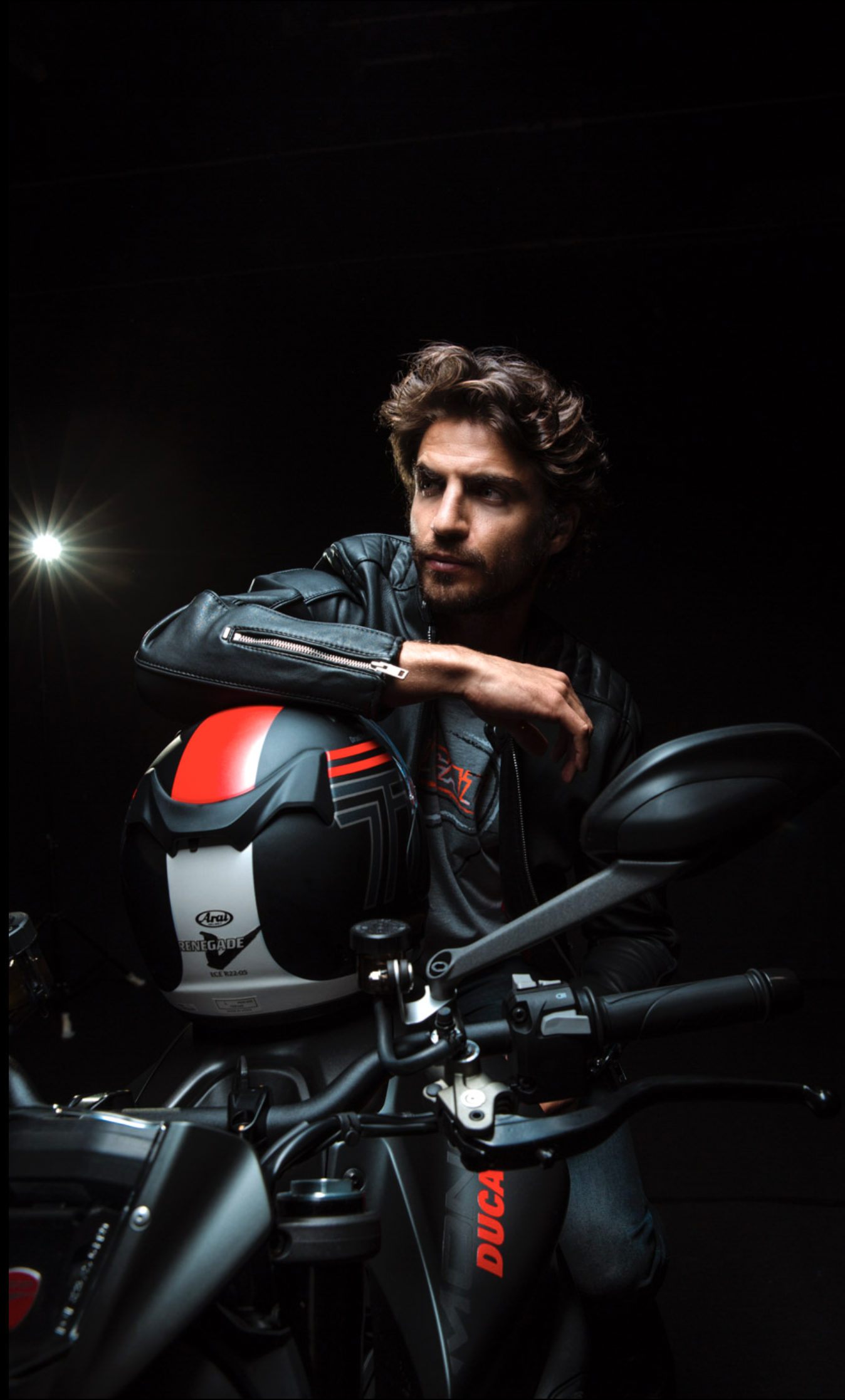 Joven con barba y cabello rizado, con chaqueta de cuero, sentado en una motocicleta negra, con casco decorado con colores negro, rojo y blanco, en un fondo oscuro y con una luz brillante al fondo a la izquierda.