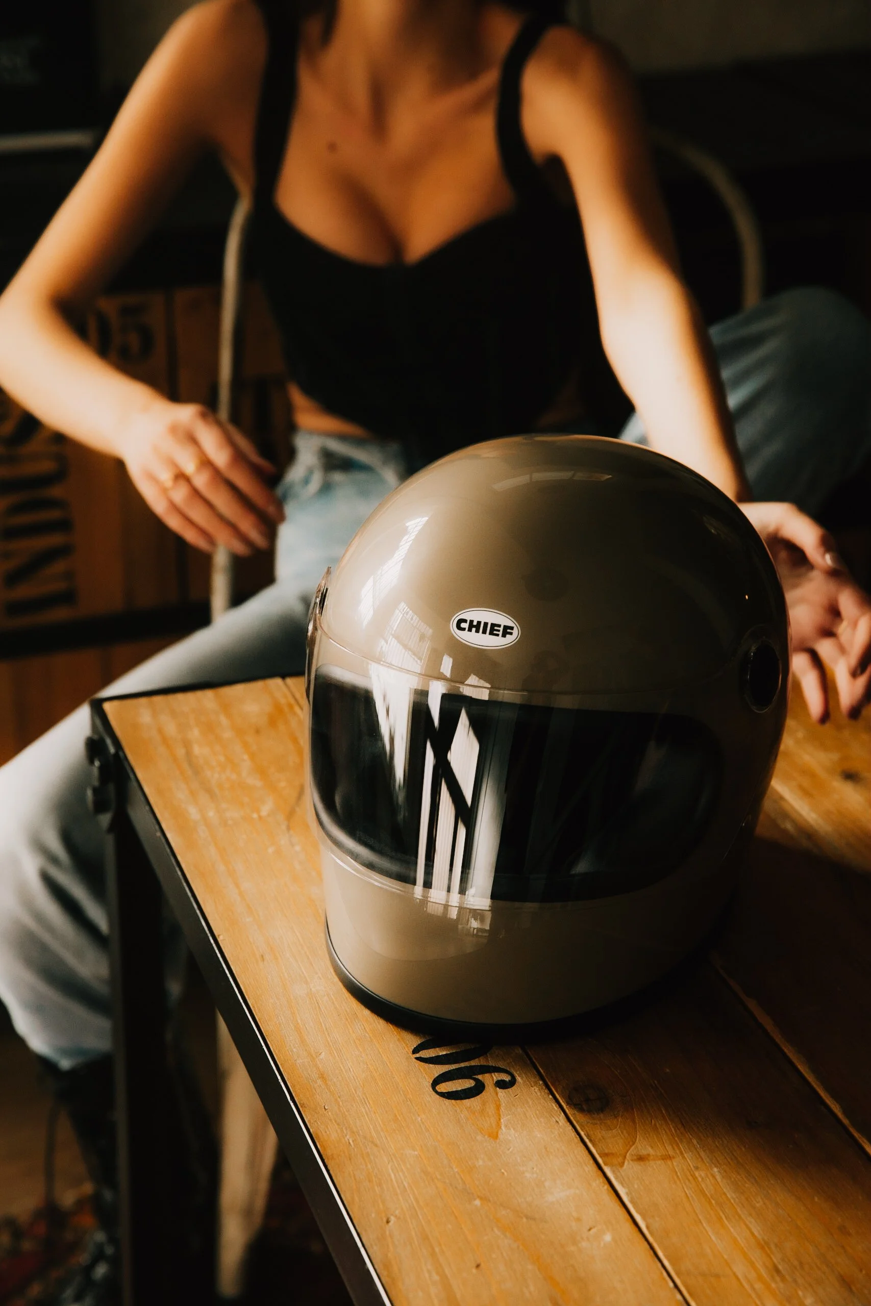 Casco de motocicleta en una mesa de madera, con una mujer sentada detrás. La mujer viste un top negro y jeans claros.