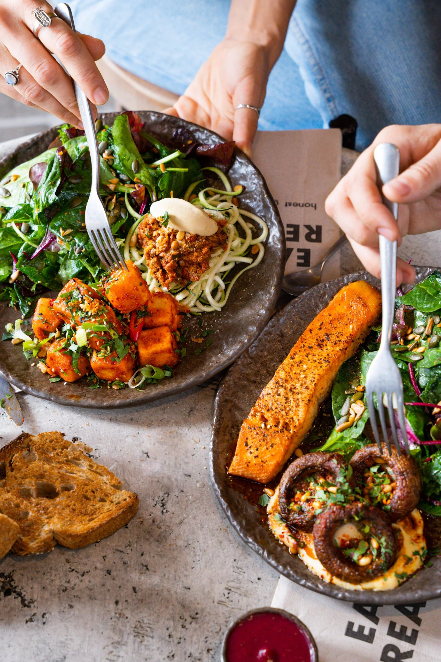 Platos de comida, incluyendo ensalada, salmón, pulpo y curry de papas, en platos de cerámica, con pan y bebida acrílica al fondo.