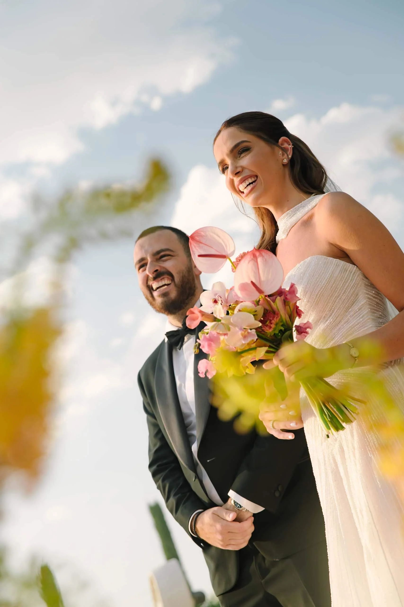 Una pareja feliz en vestido y esmoquin en exterior con cielo despejado, ella sostiene un ramo de flores rosadas y él entrelaza sus manos.