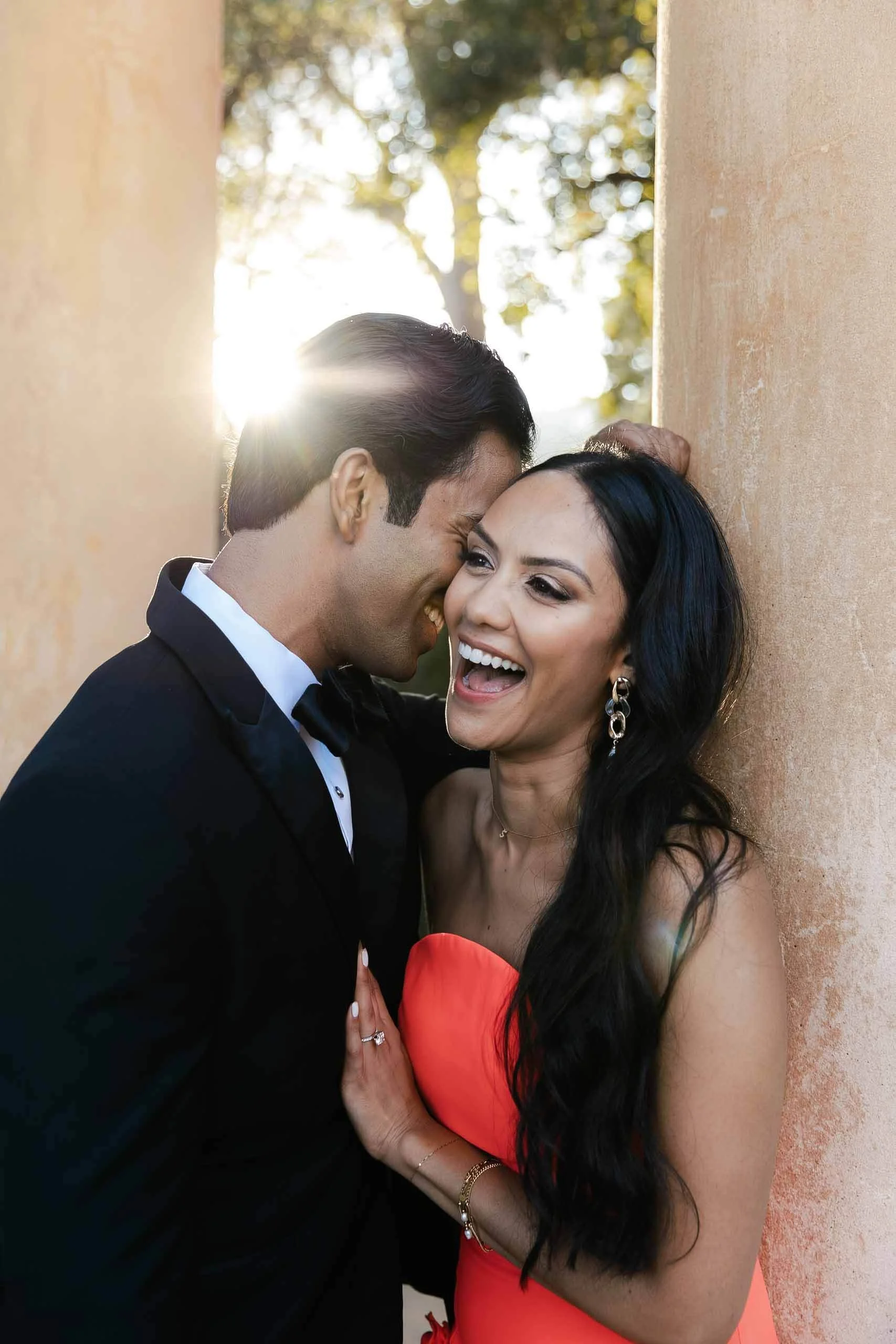 Una pareja de adultos jóvenes riendo y abrazados, la mujer lleva un vestido rojo y el hombre usa traje negro con moño, están apoyados en una pared de color terracota con fondo de árboles y luz del sol