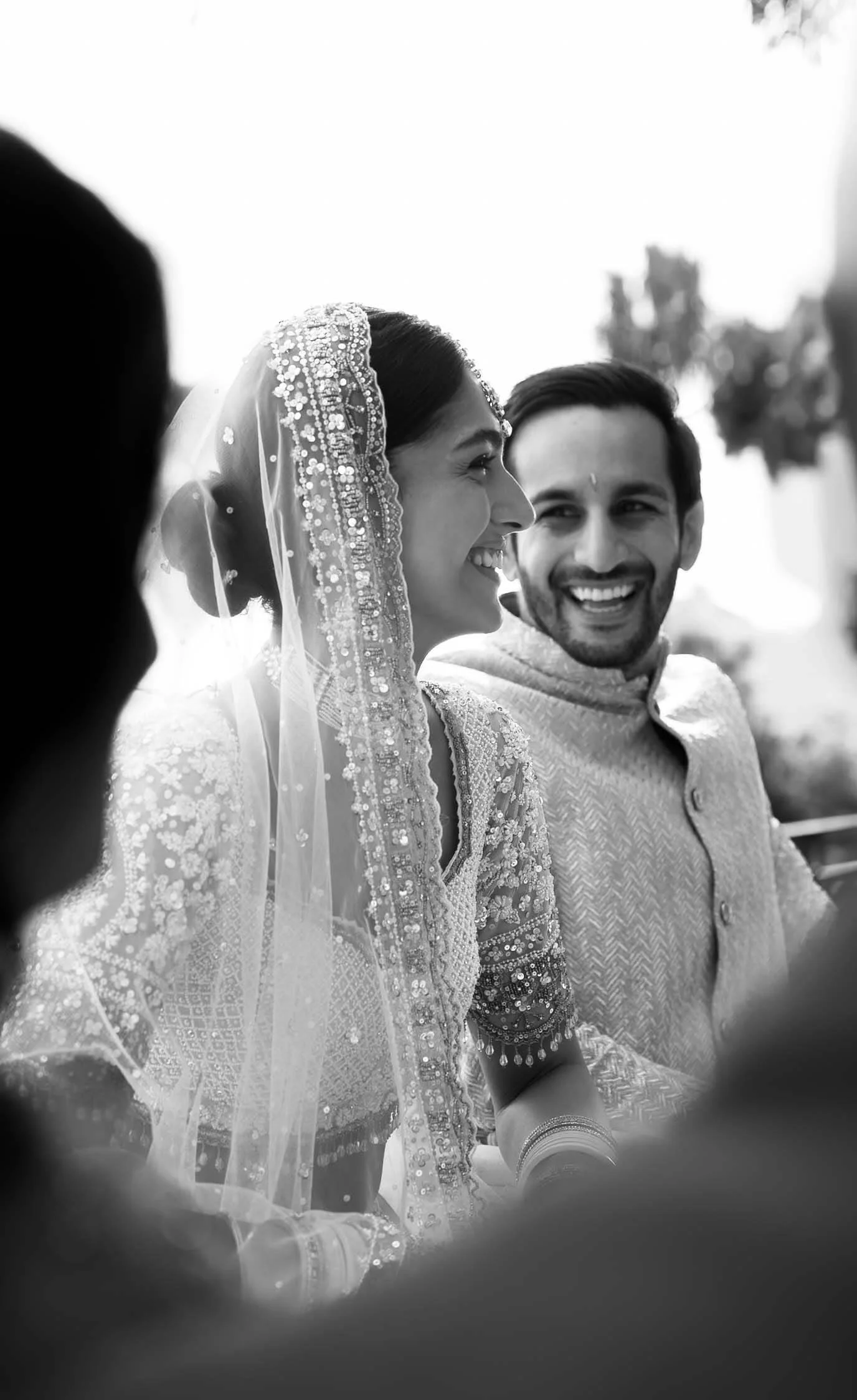 Imagen en blanco y negro de una pareja en su boda, la mujer lleva un vestido con detalles de lentejuelas y un velo adornado con perlas y pedrería, ambos parecen felices y compartiendo un momento especial.