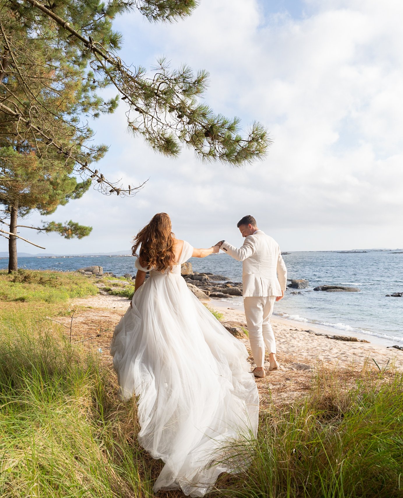 Una pareja en un matrimonio en la playa, la mujer en vestido de novia y el hombre en traje blanco, sosteniéndose de las manos mientras caminan hacia el mar.
