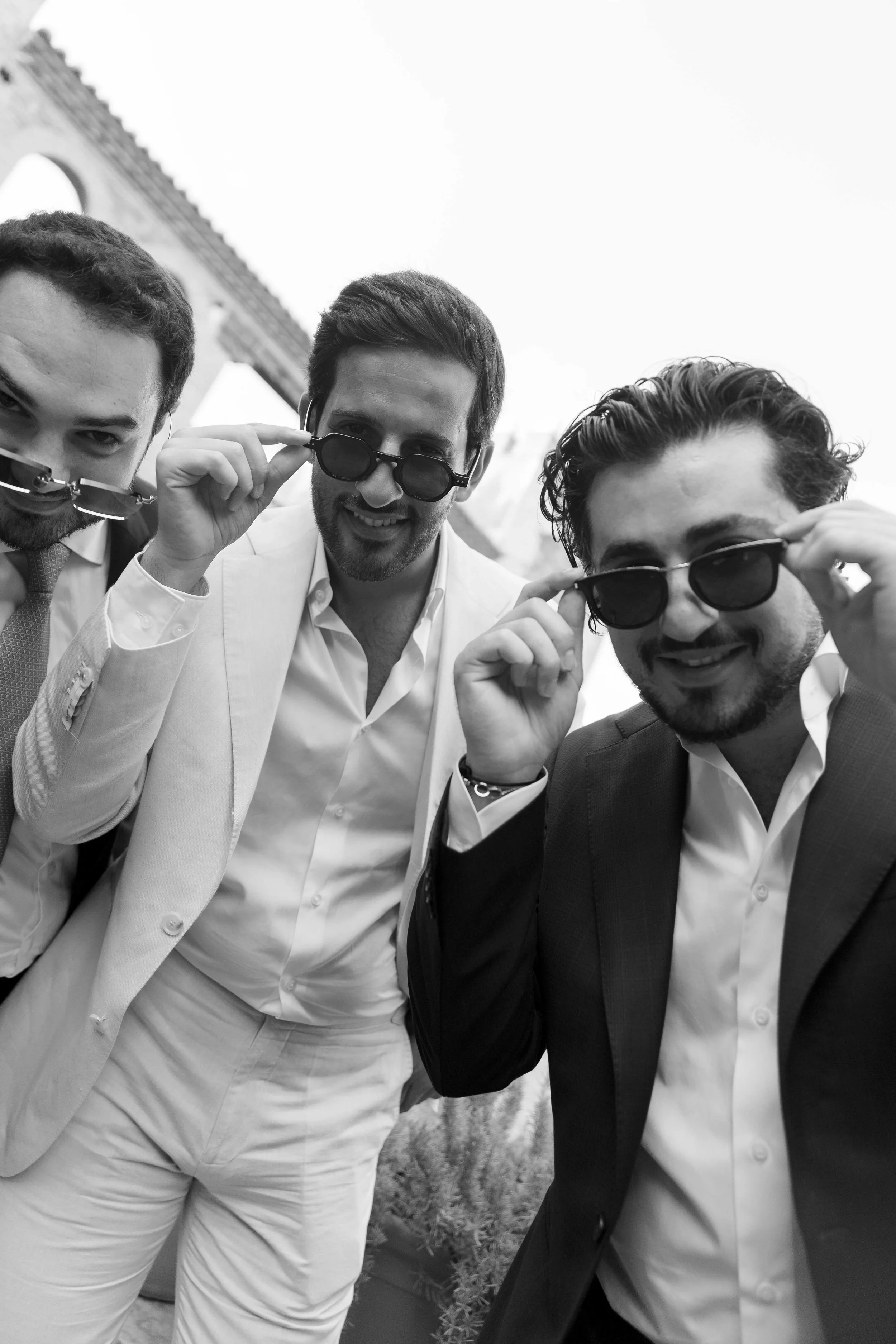 Tres hombres con trajes y gafas de sol posando para la foto en un lugar alto con estructura arquitectónica visible y cielo despejado.