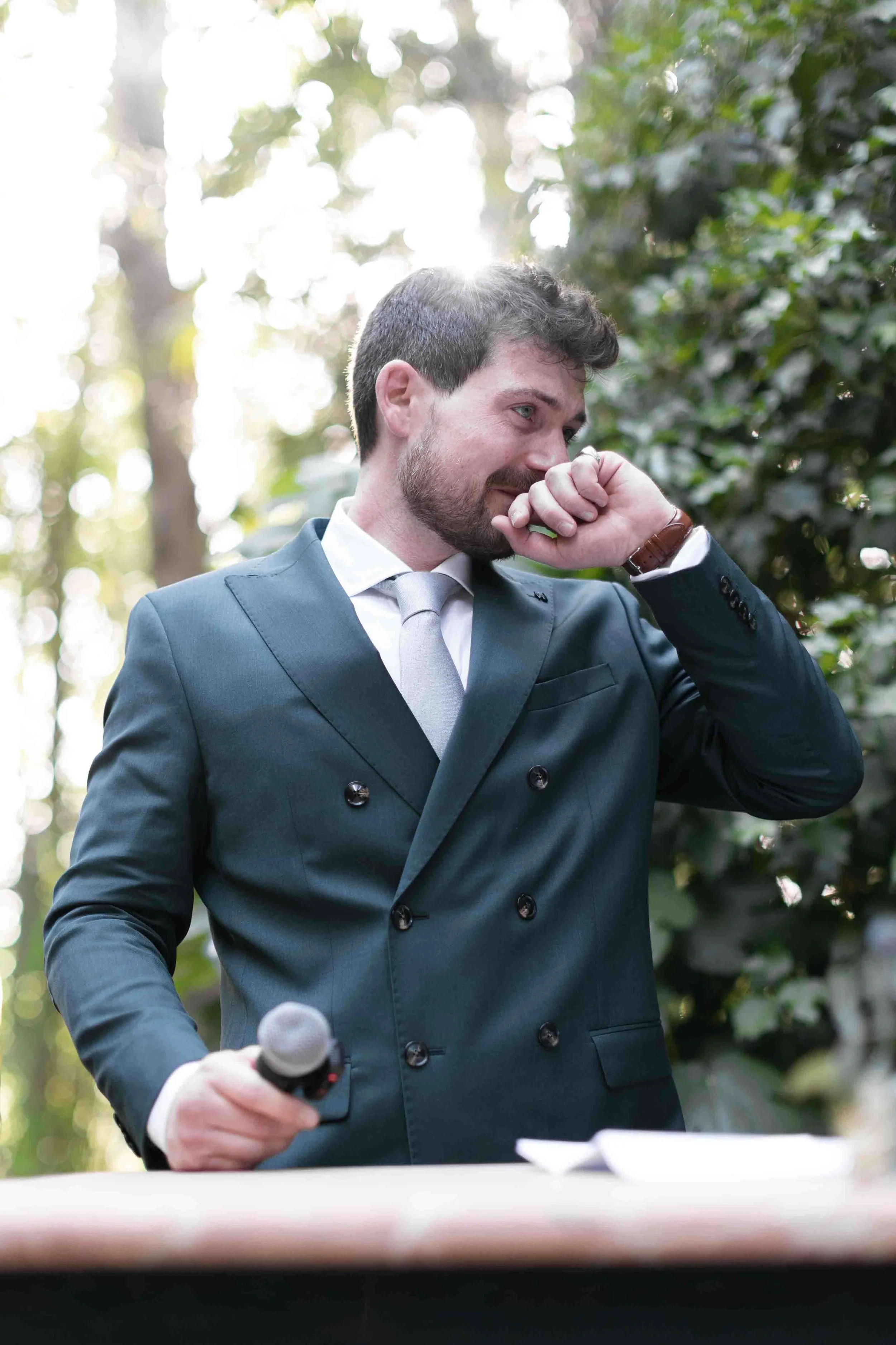 Hombre de traje azul oscuro y corbata gris, sosteniendo un micrófono, en una reunión al aire libre, rodeado de árboles y con luz solar de fondo.