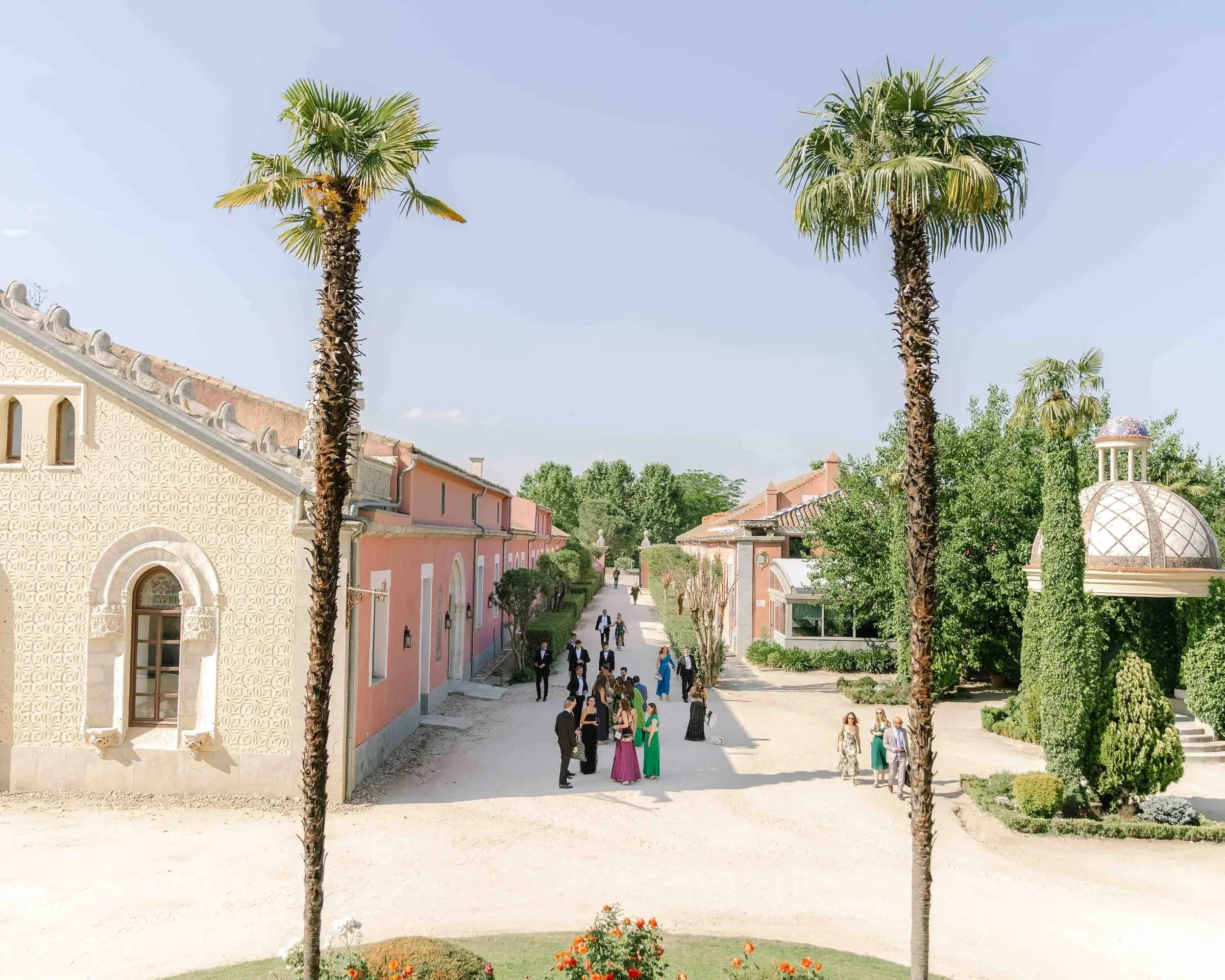 Palacio de viñuelas en Vista de un evento al aire libre con personas vestidas de noche, en un espacio con palmeras, árboles, edificios de colores suaves y un cielo despejado.